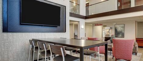 A modern hotel lobby with a dining area, a flat-screen TV mounted on the wall, and a 'The Market' sign visible in the background.