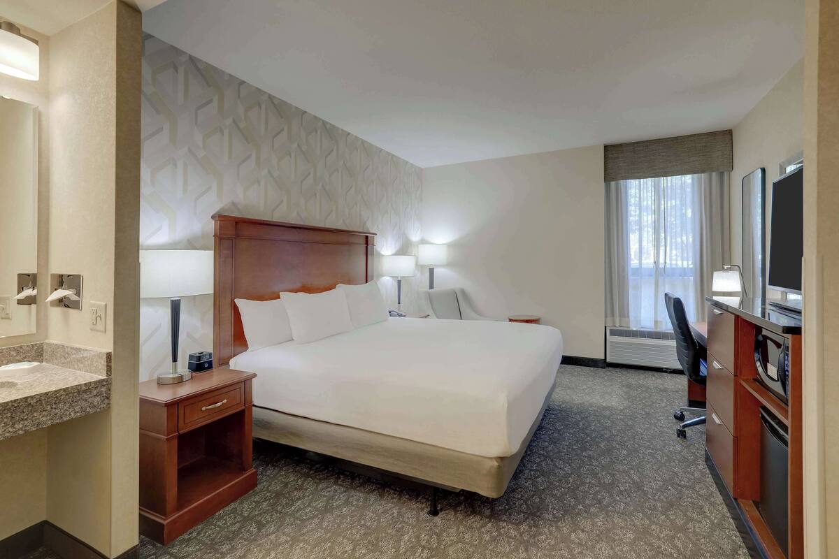 A hotel room with a large bed, a desk with a chair, a vanity with a mirror, and a window with curtains.