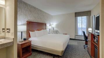 A hotel room with a large bed, a desk with a chair, a vanity with a mirror, and a window with curtains.
