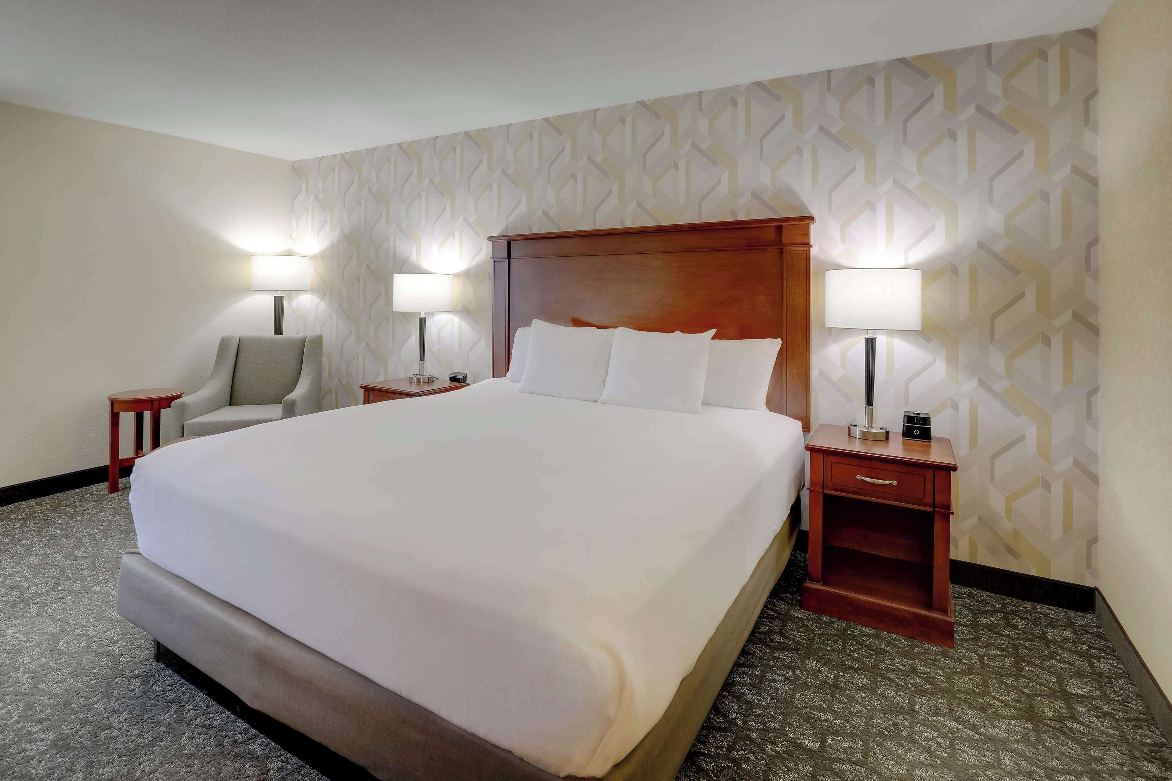A hotel room with a large bed, two bedside tables with lamps, a chair, and a patterned wall.