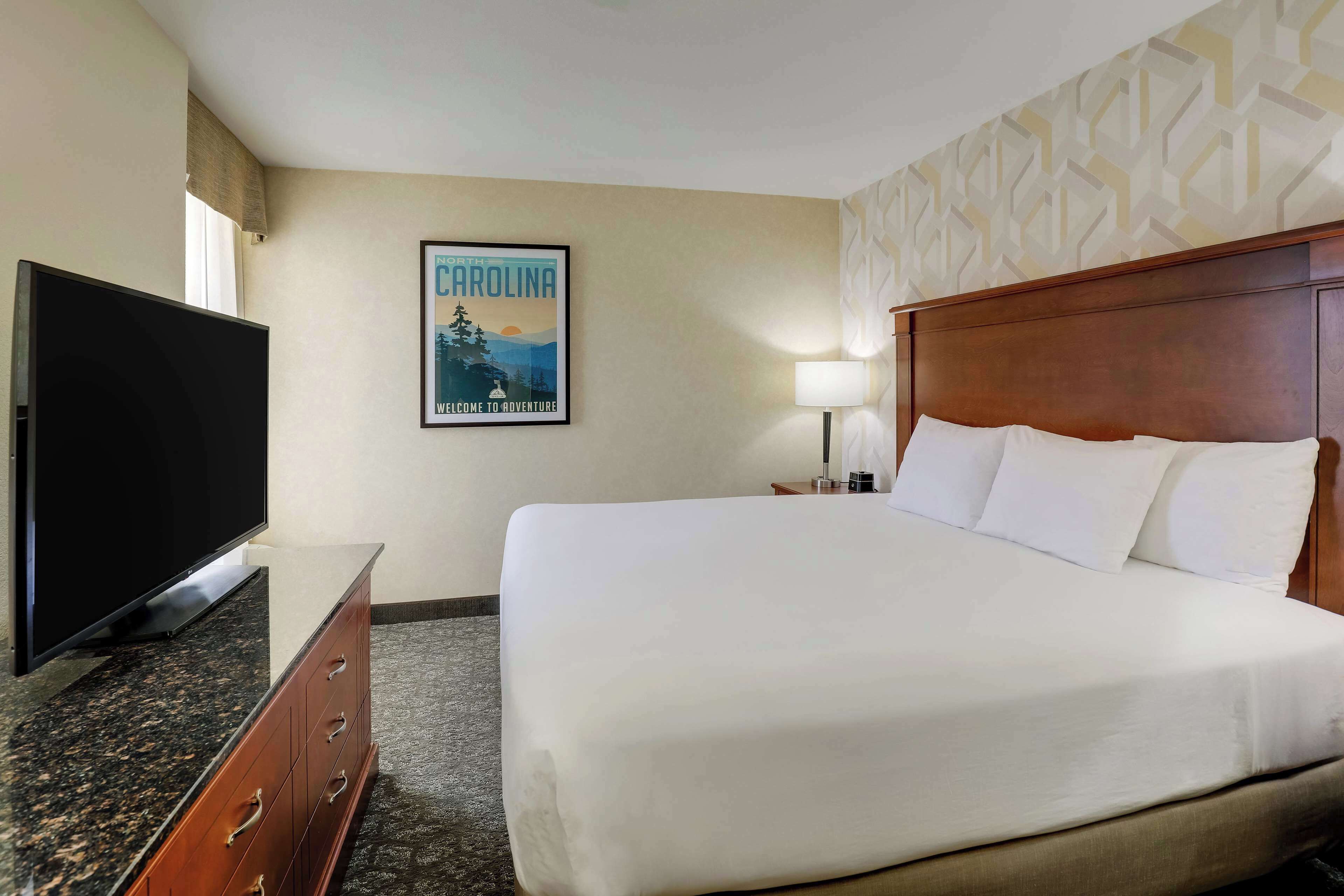 Une chambre d’hôtel avec un grand lit, un téléviseur à écran plat, une commode en bois et une affiche de bienvenue de la Caroline du Nord.