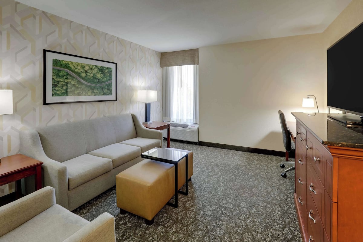 A hotel room with a sofa, ottoman, small table, desk, TV, and a window with curtains.