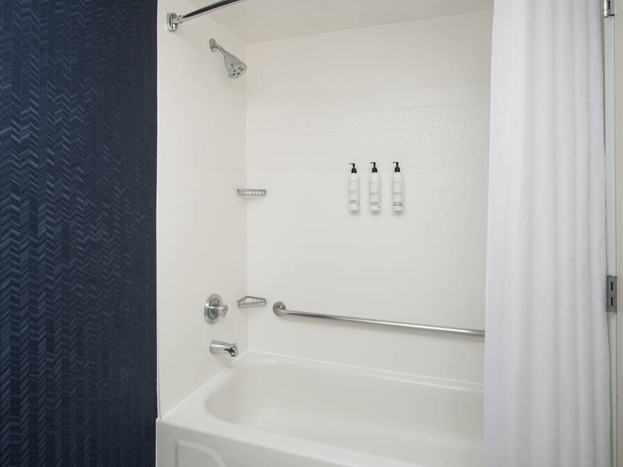Un baño con una bañera blanca, un cabezal de ducha, un pasamanos y tres botellas de champú.