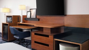 Ett hotellrum med ett skrivbord i trä, en platt-TV, en lampa, en stol, ett litet skåp och en papperskorg.