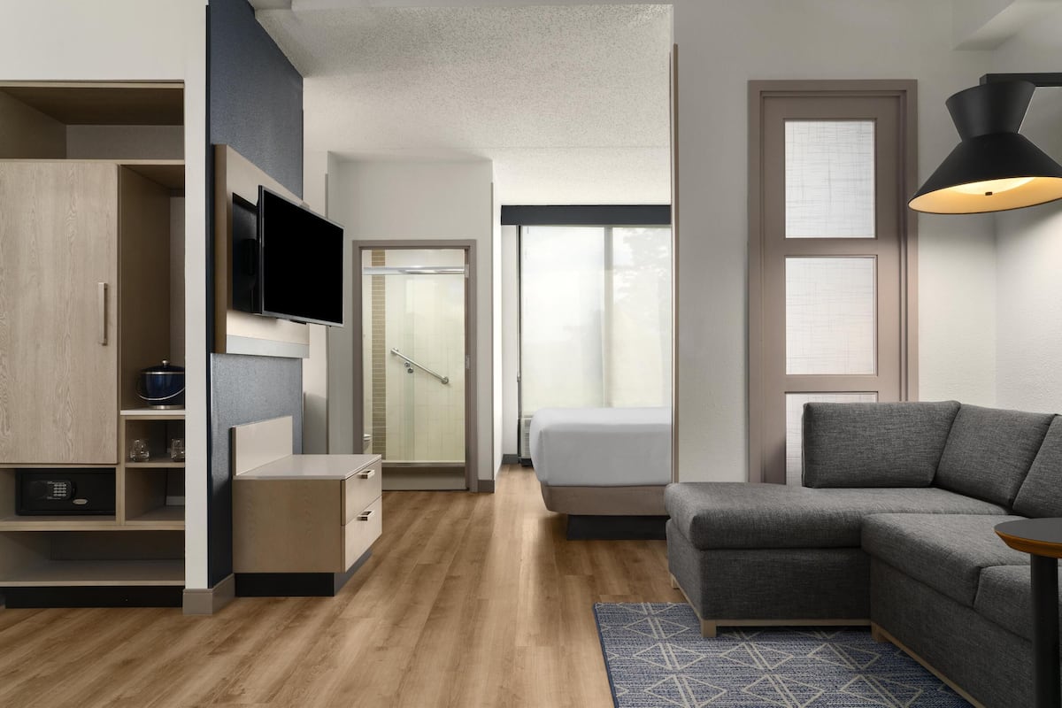 Une chambre d’hôtel moderne avec un canapé gris, un téléviseur à écran plat, un dressing et un espace douche.