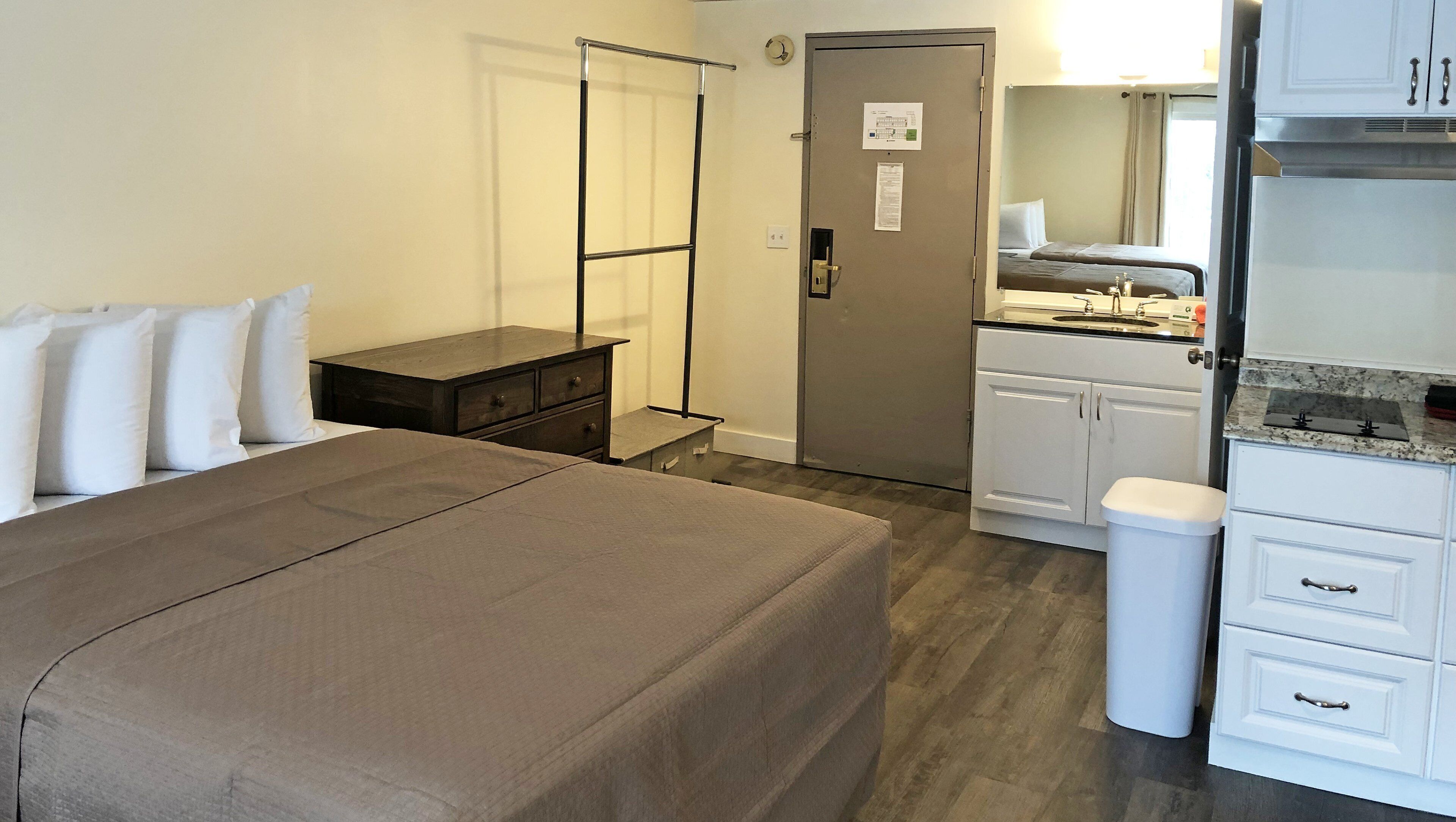 Habitación de hotel con cama, tocador pequeño, puerta, zona de cocina con refrigerador y cubo de basura.