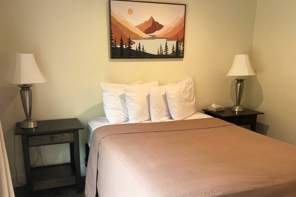 Un lit bien fait, avec du linge de lit blanc et une couvre-lit marron, est encadré de deux tables de chevet sur lesquelles sont posées des lampes. L’une des tables de chevet porte un boîte de mouchoirs. Un tableau représentant un paysage, encadré, est accroché au-dessus du lit.
