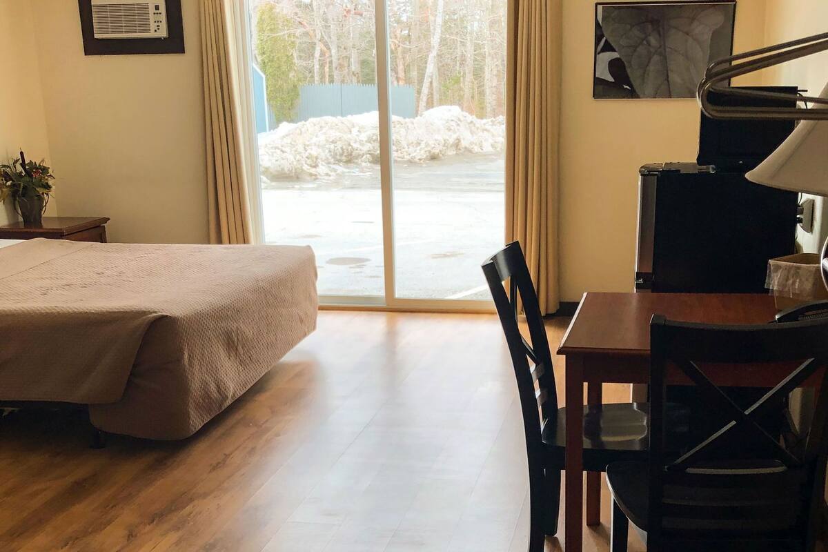 Une chambre d’hôtel avec un lit, un bureau, une chaise, un piano et une grande fenêtre donnant sur des arbres et une clôture.