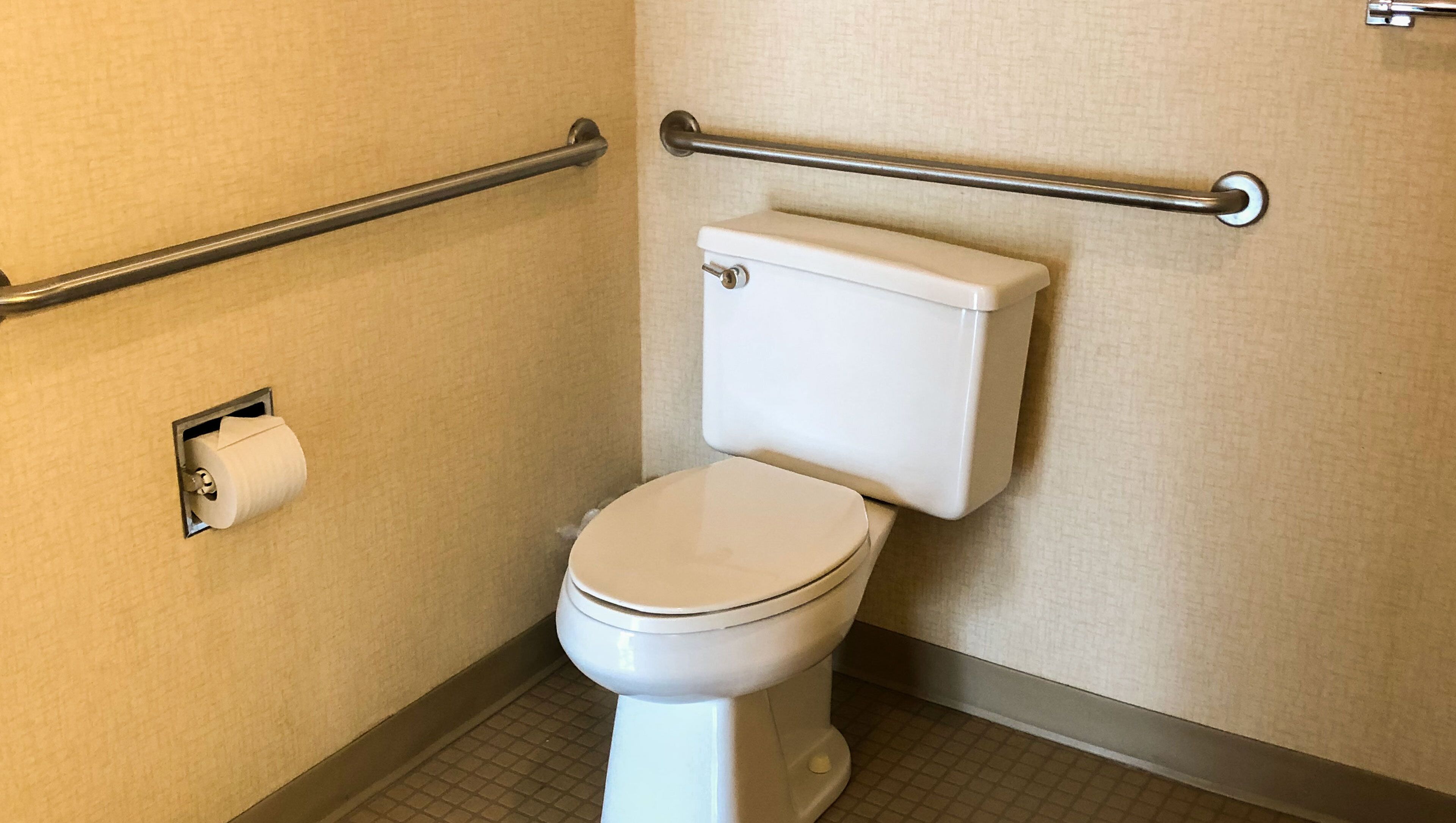 A standard toilet with a white tank and seat, a paper towel dispenser, and a grab bar on the wall.