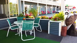 A motel with a patio area featuring green chairs and a table, surrounded by flower beds and a bird sculpture.