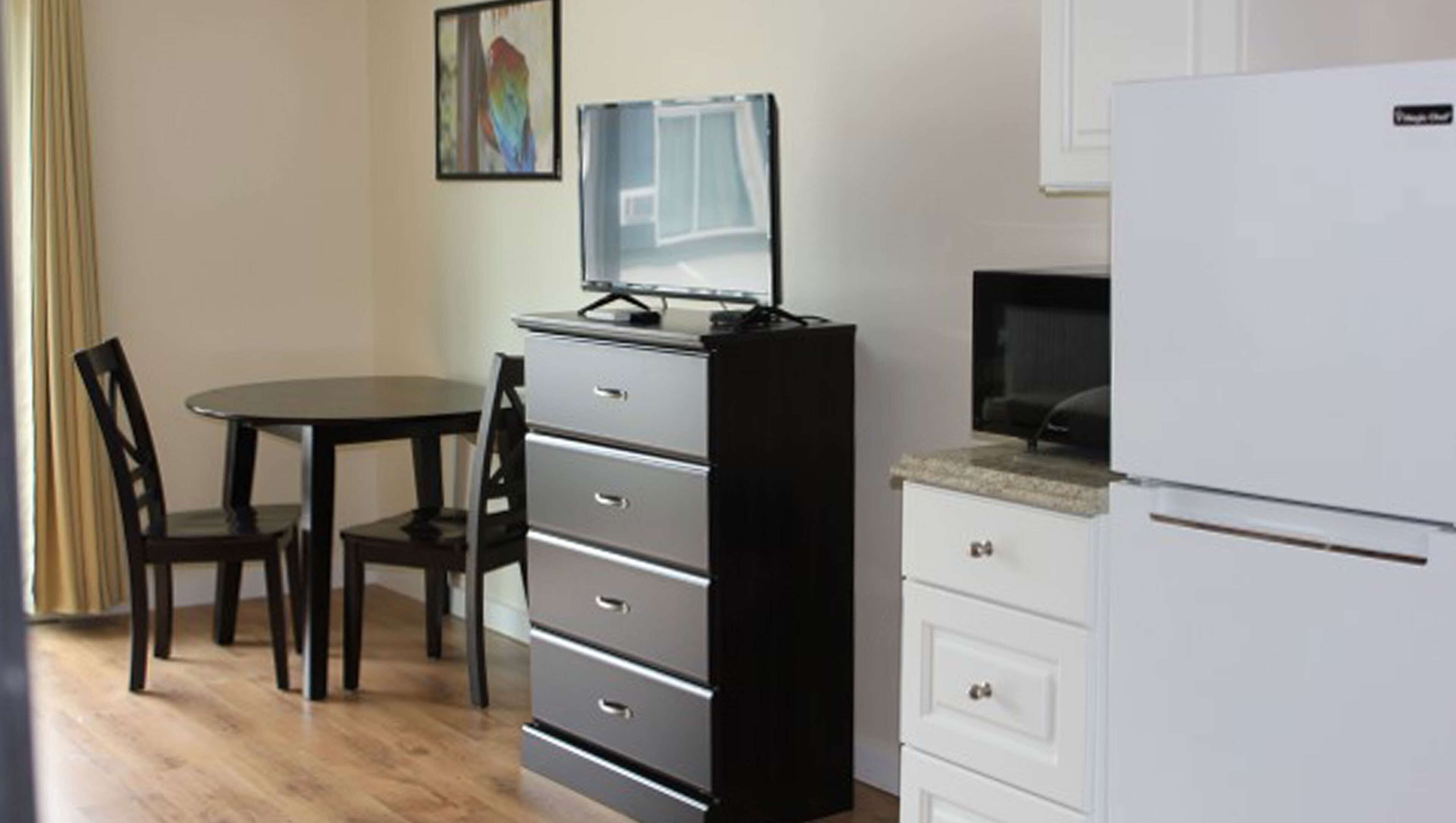 Una habitación pequeña con mesa de comedor, dos sillas, un archivador negro, un refrigerador blanco y un televisor sobre un soporte.