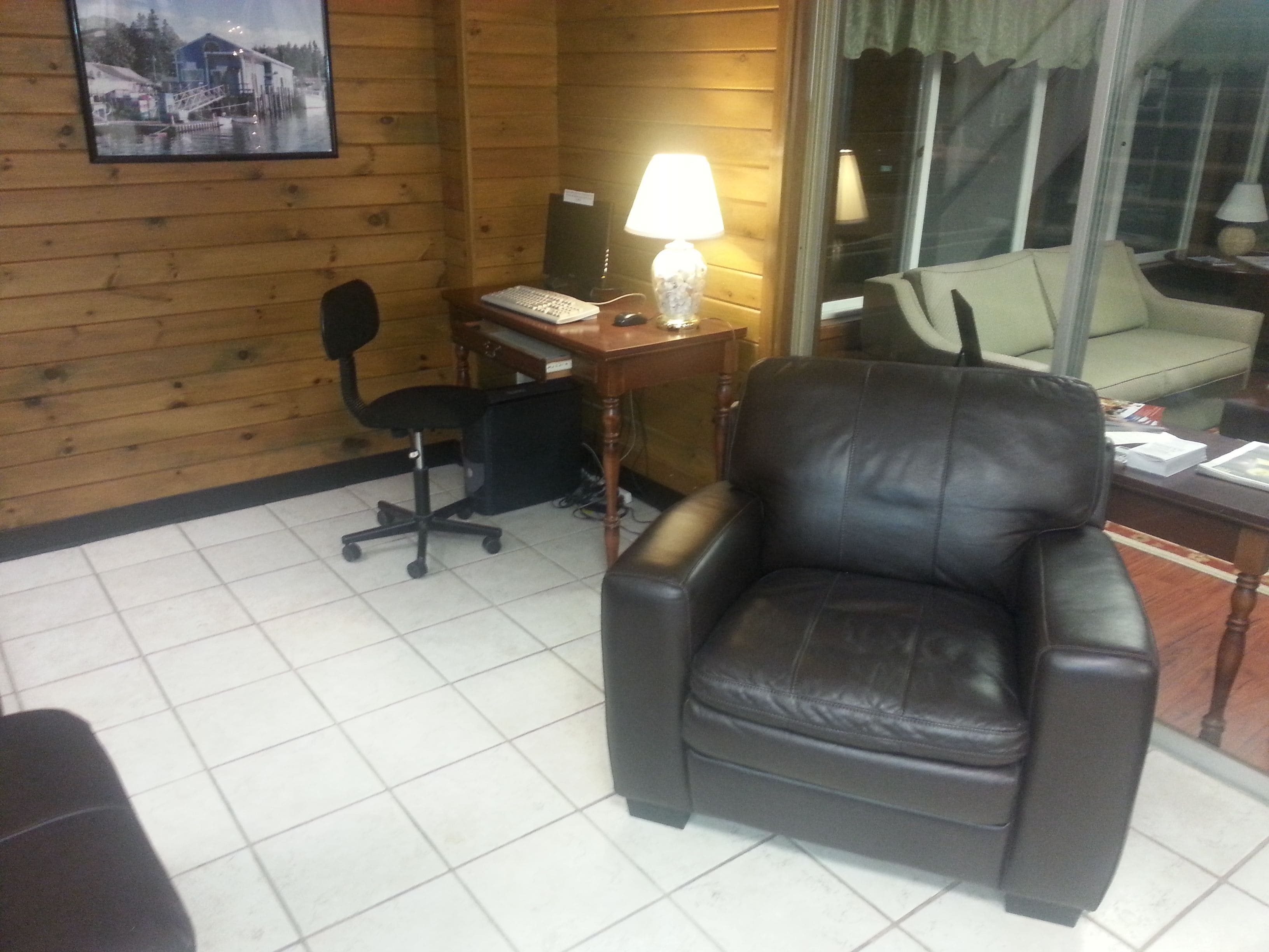 Una habitación con un sillón de cuero, un escritorio con computadora y lámpara, una silla y una ventana con vista a un edificio.