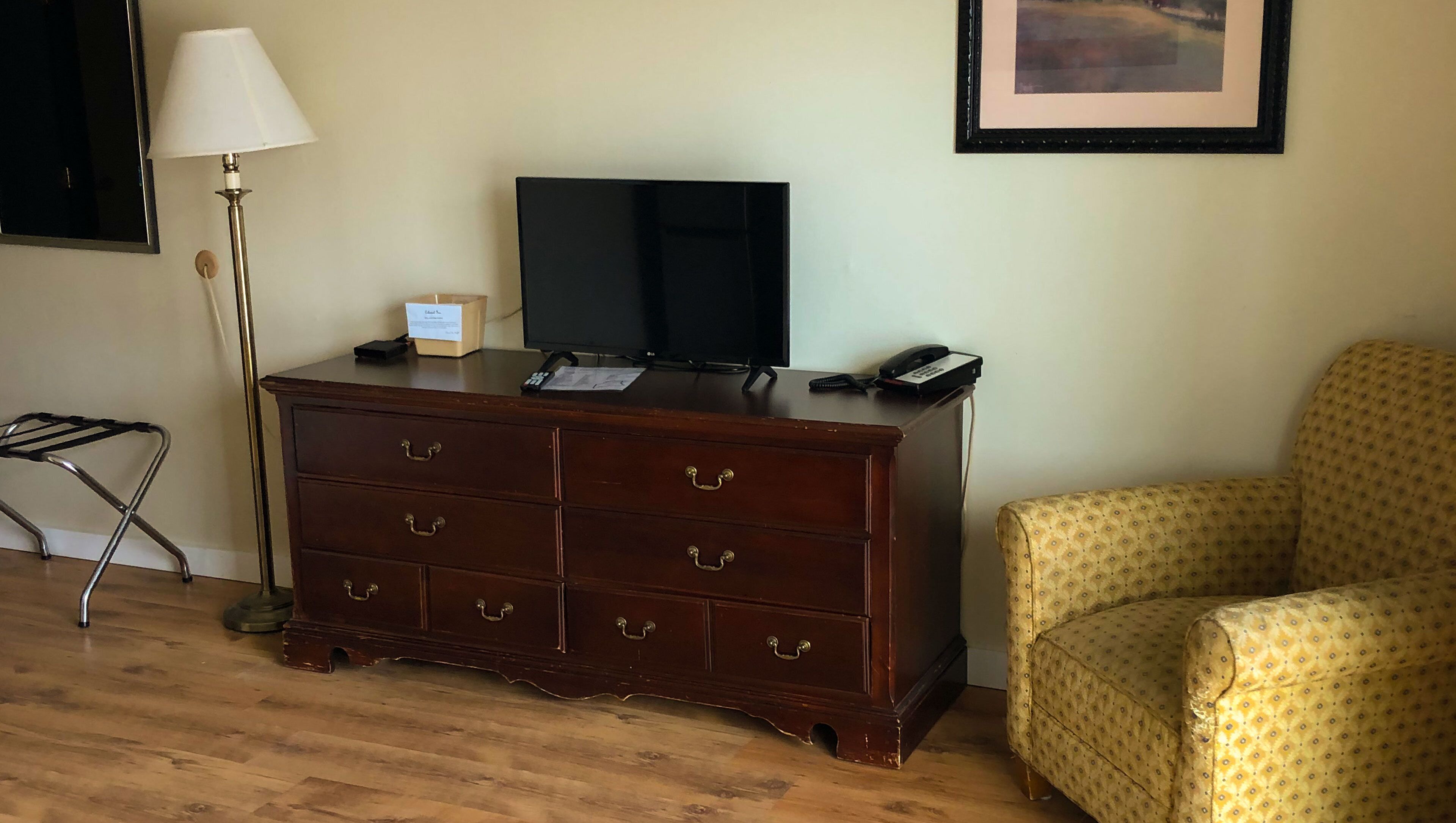 A room with a wooden dresser, a television, a lamp, a chair, and a framed picture on the wall.