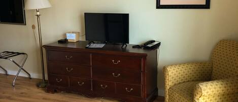 A room with a wooden dresser, a television, a lamp, a chair, and a framed picture on the wall.