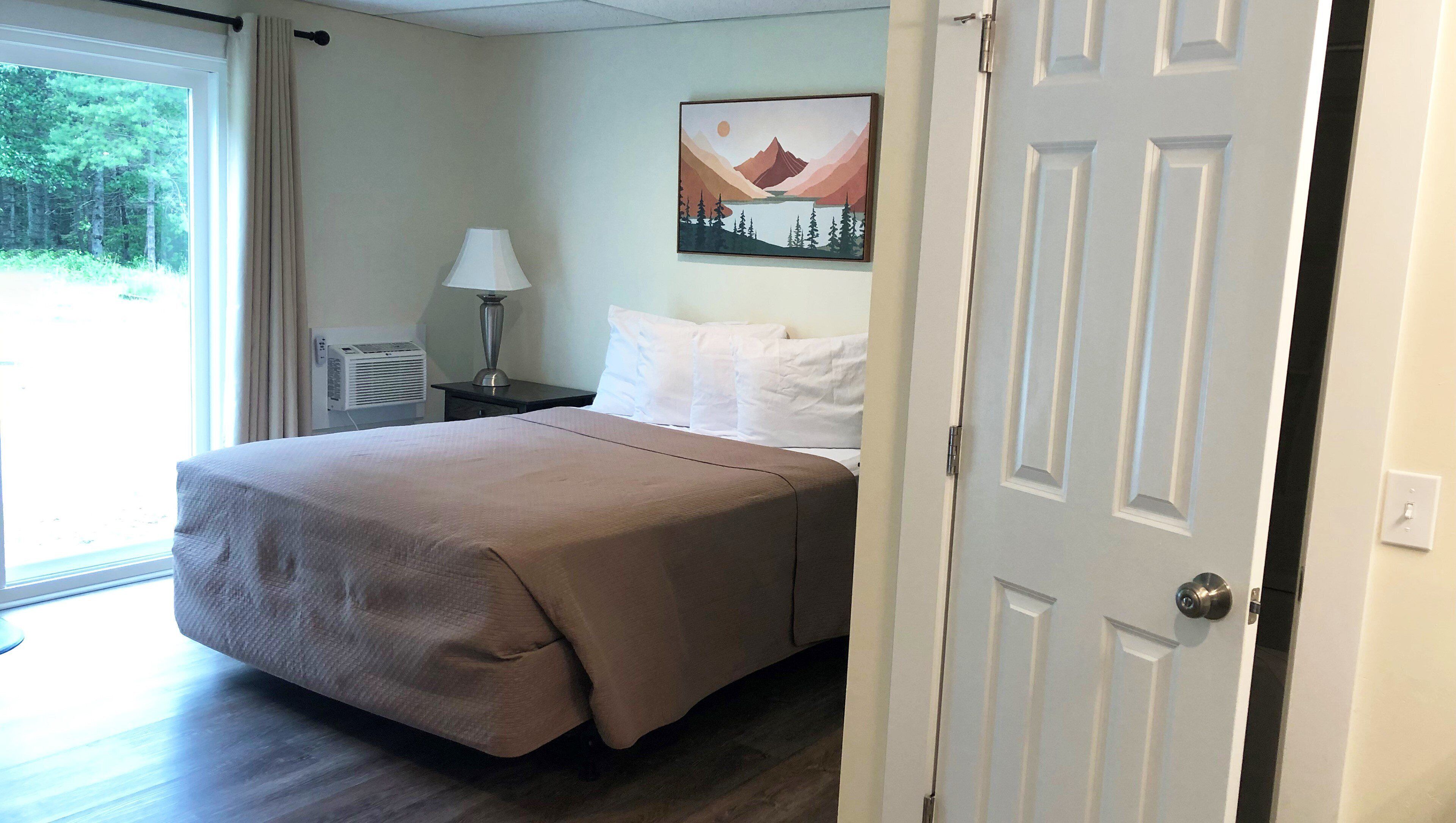 A hotel room with a bed, a nightstand, a lamp, a painting, and a door.