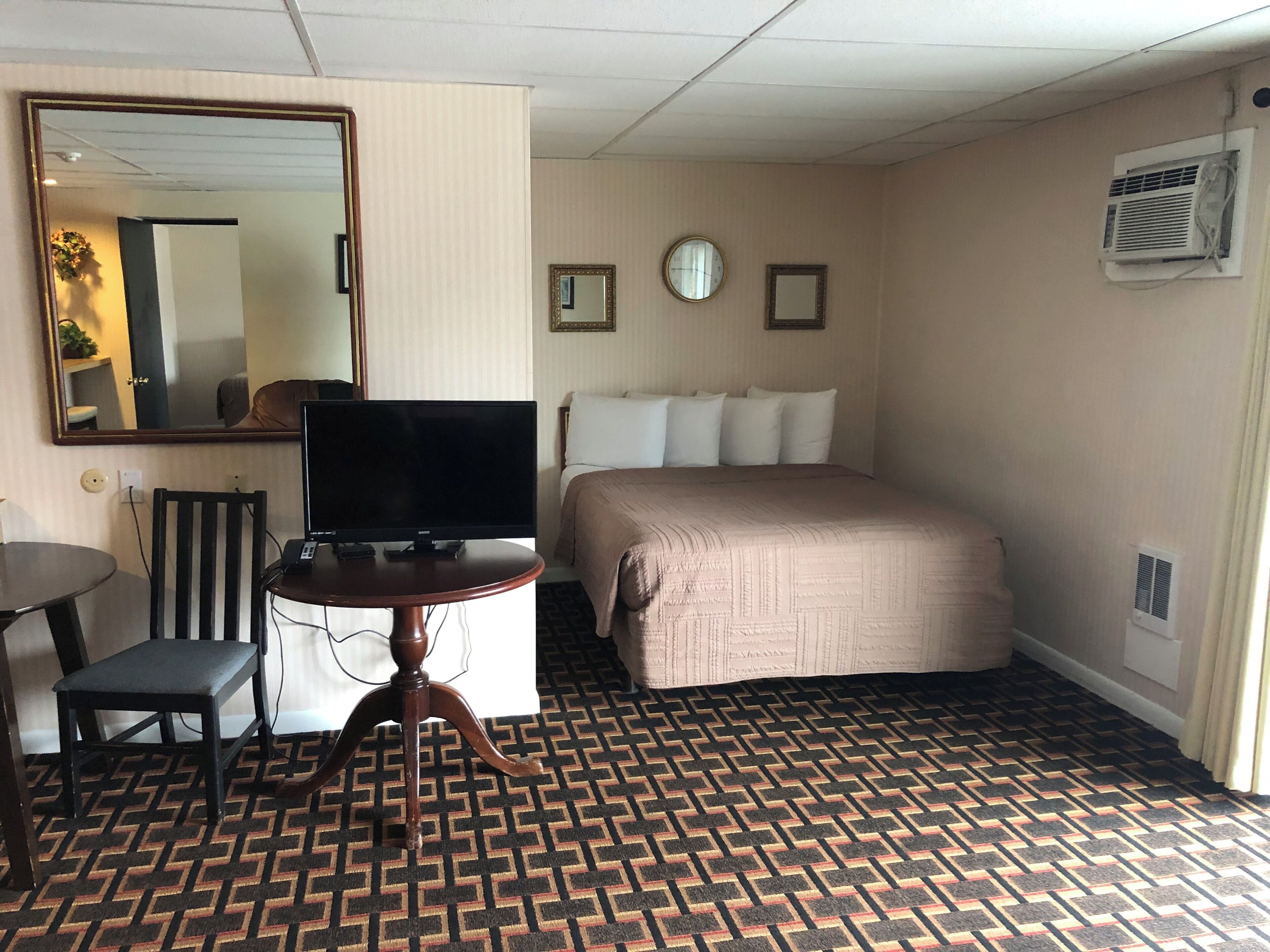 A hotel room with a bed, a television on a wooden table, a mirror, and a patterned carpet.