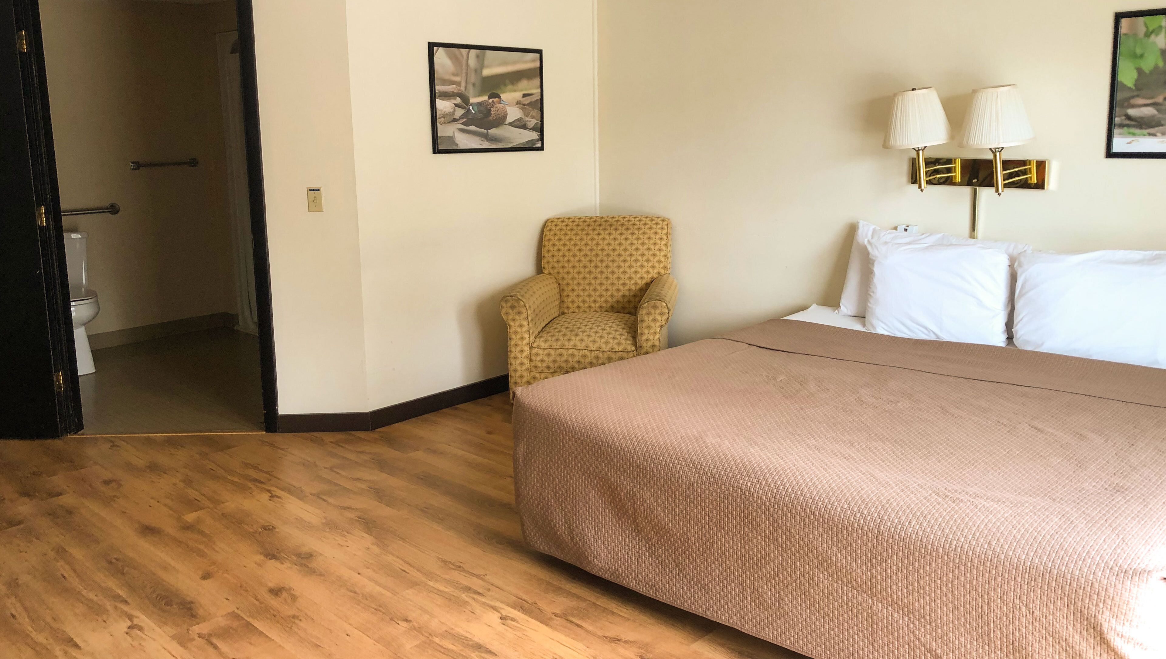 A hotel room with a wooden floor, a bed with a beige bedspread, a patterned armchair, a white toilet, and a framed picture on the wall.