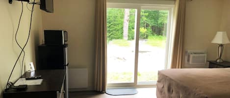 Habitación de hotel con cama, escritorio con televisor, lámpara y vista a un bosque desde la puerta.