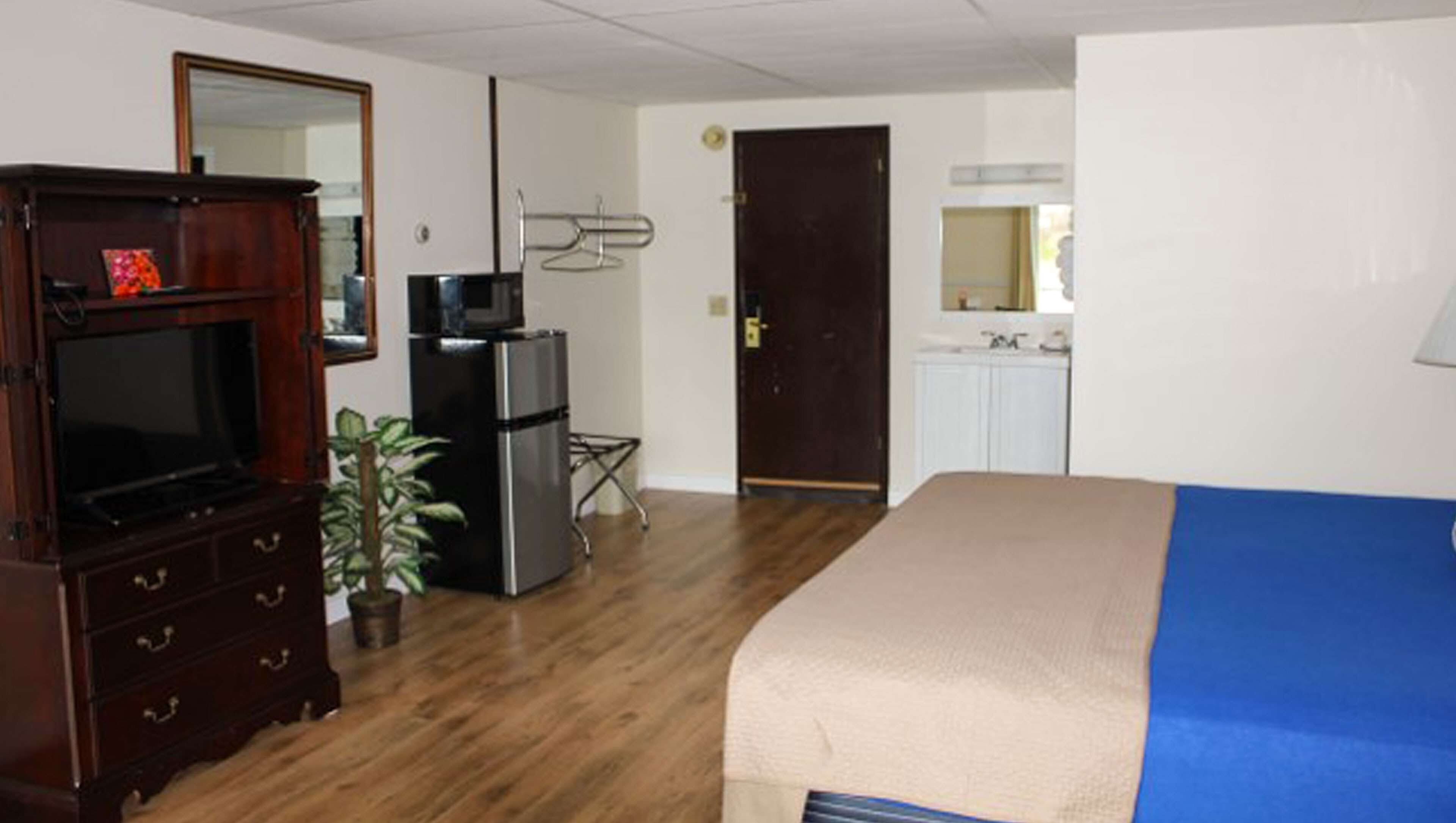 A hotel room with a wooden dresser, a television, a refrigerator, a microwave, a bed with a blue blanket, a potted plant, and a door leading to a bathroom.