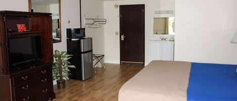 A hotel room with a wooden dresser, a television, a refrigerator, a microwave, a bed with a blue blanket, a potted plant, and a door leading to a bathroom.
