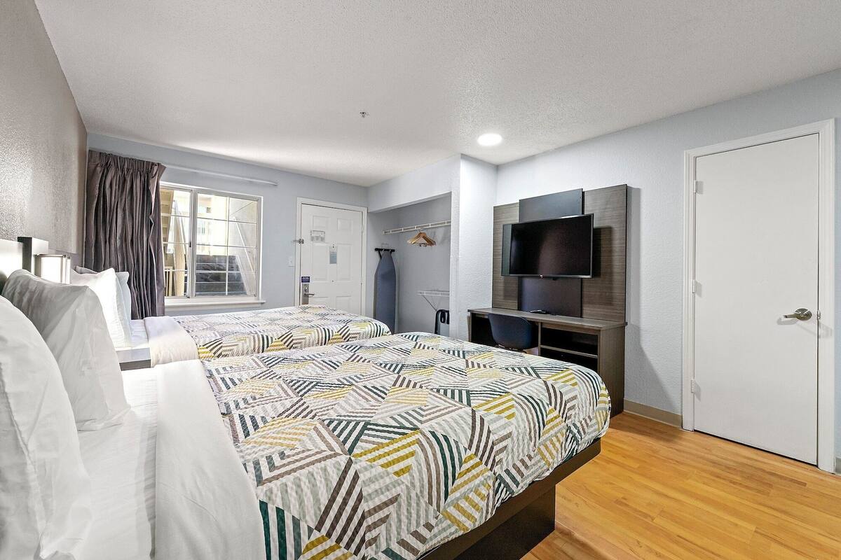 A hotel room with two beds, a TV, a desk, and a window with curtains.