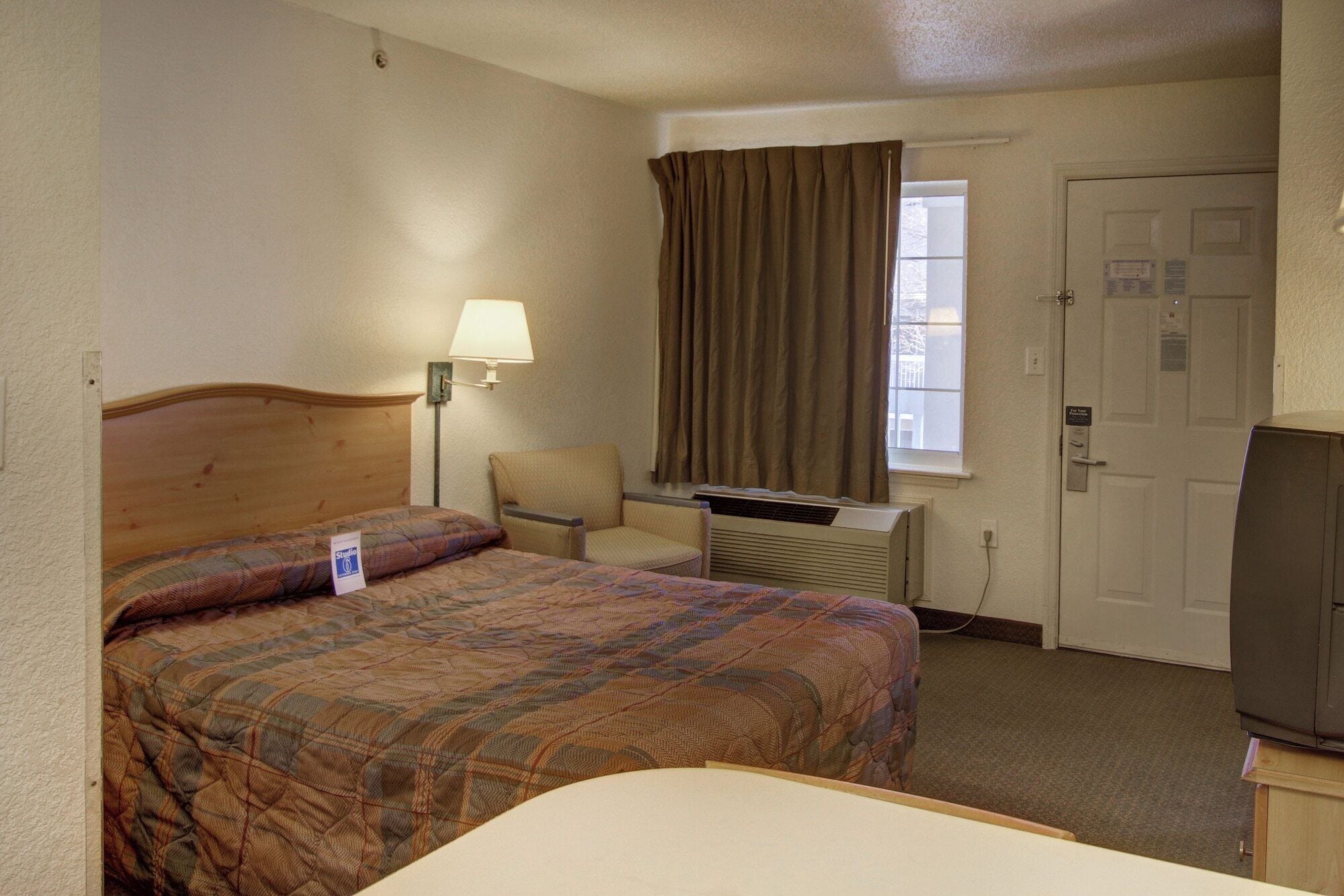 Quarto de hotel com cama, cadeira, escrivaninha, televisão e janela com cortinas.