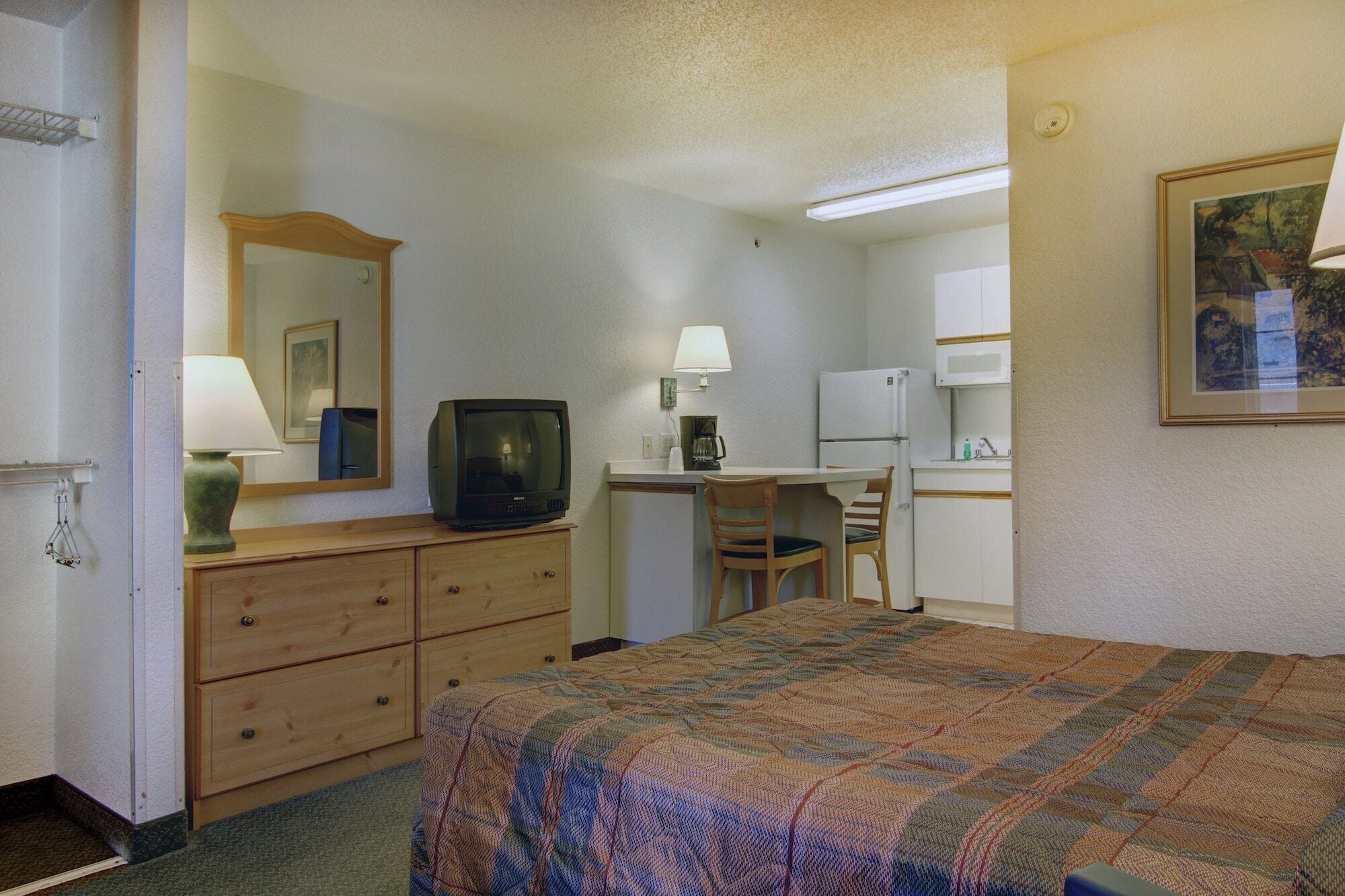 A compact hotel room with a bed, a desk, a chair, a television, a refrigerator, and a microwave.