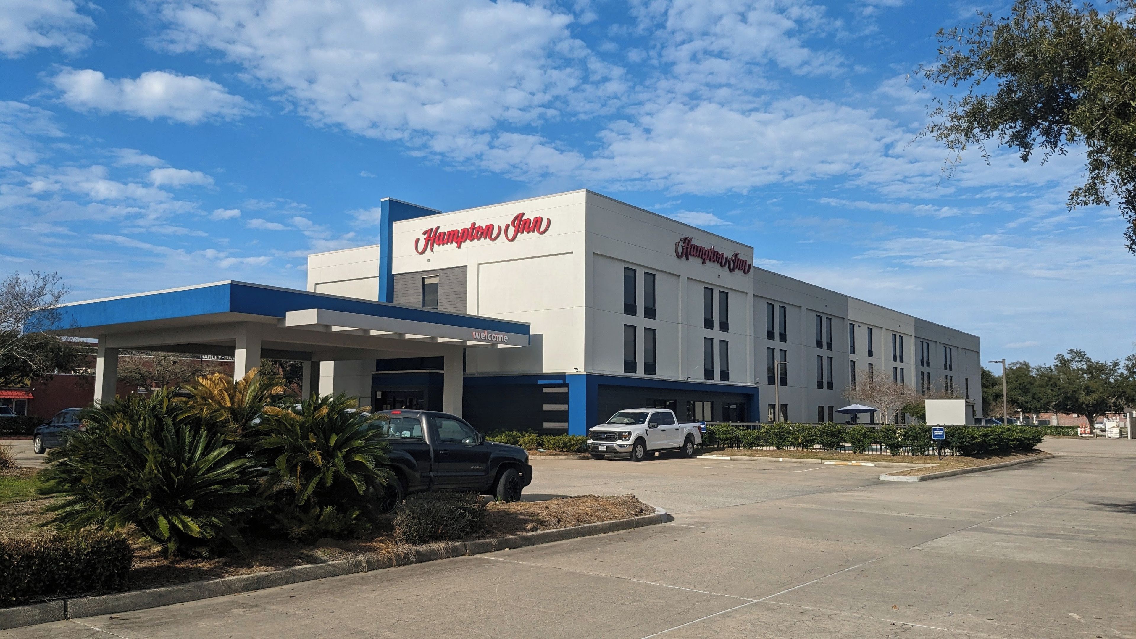 Un hotel Hampton Inn con fachada azul y blanca, zona de estacionamiento techada y cielo despejado.