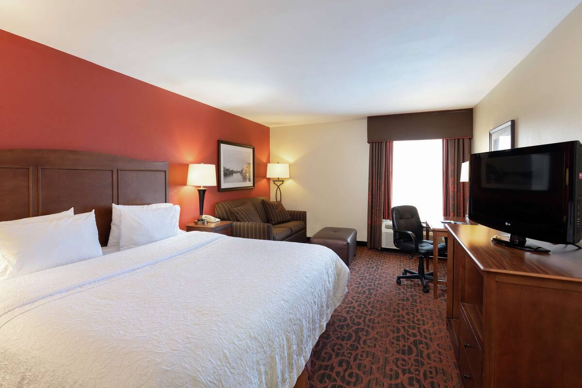 A hotel room with a large bed, a desk with a television, a chair, a sofa, and a window with curtains.