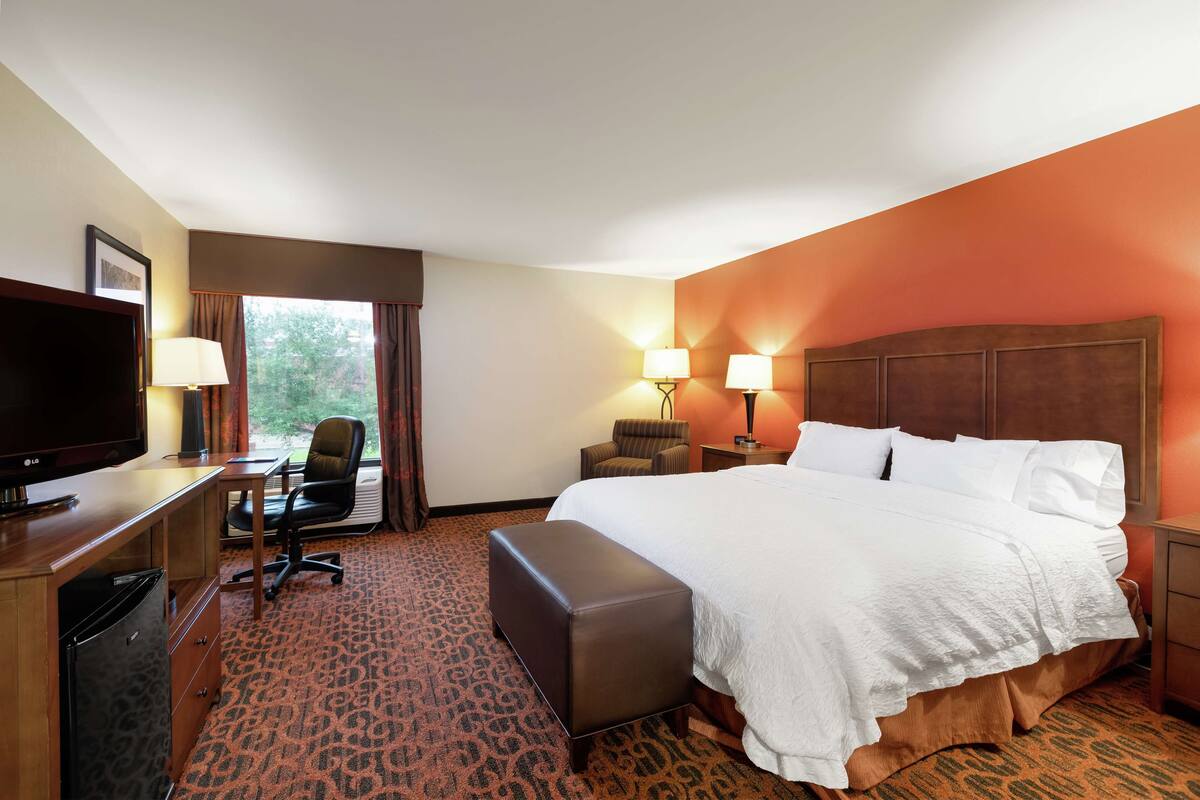 A hotel room with a bed, a desk with a television, a chair, two lamps, and a window with curtains.