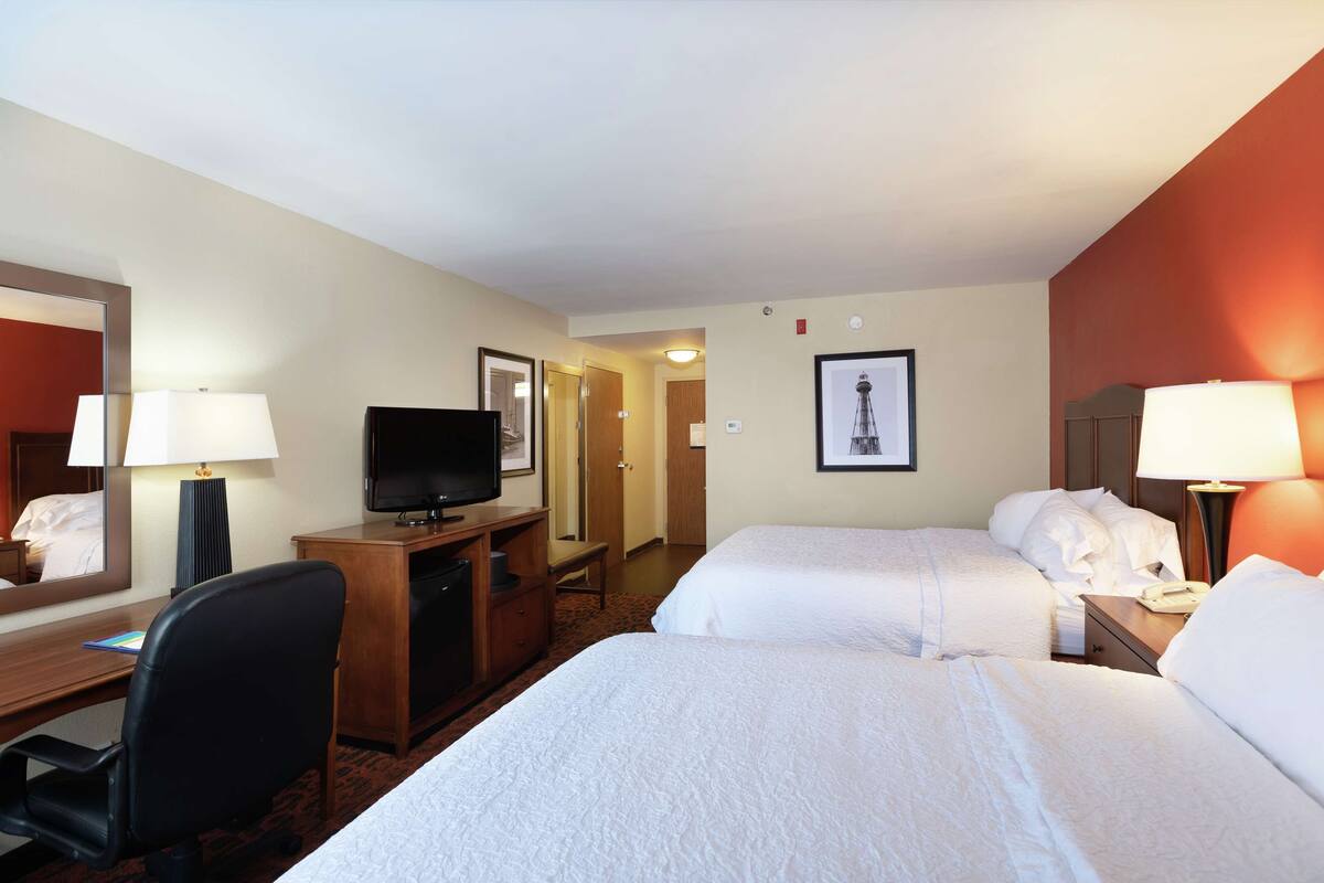 A hotel room with two beds, a desk with a TV, a mirror, and a picture of a lighthouse.