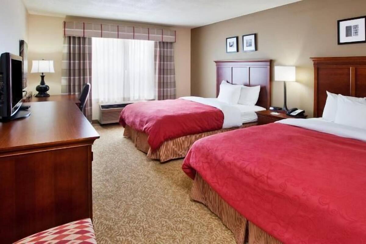A hotel room with two beds, a desk with a computer, a lamp, and a window with curtains.