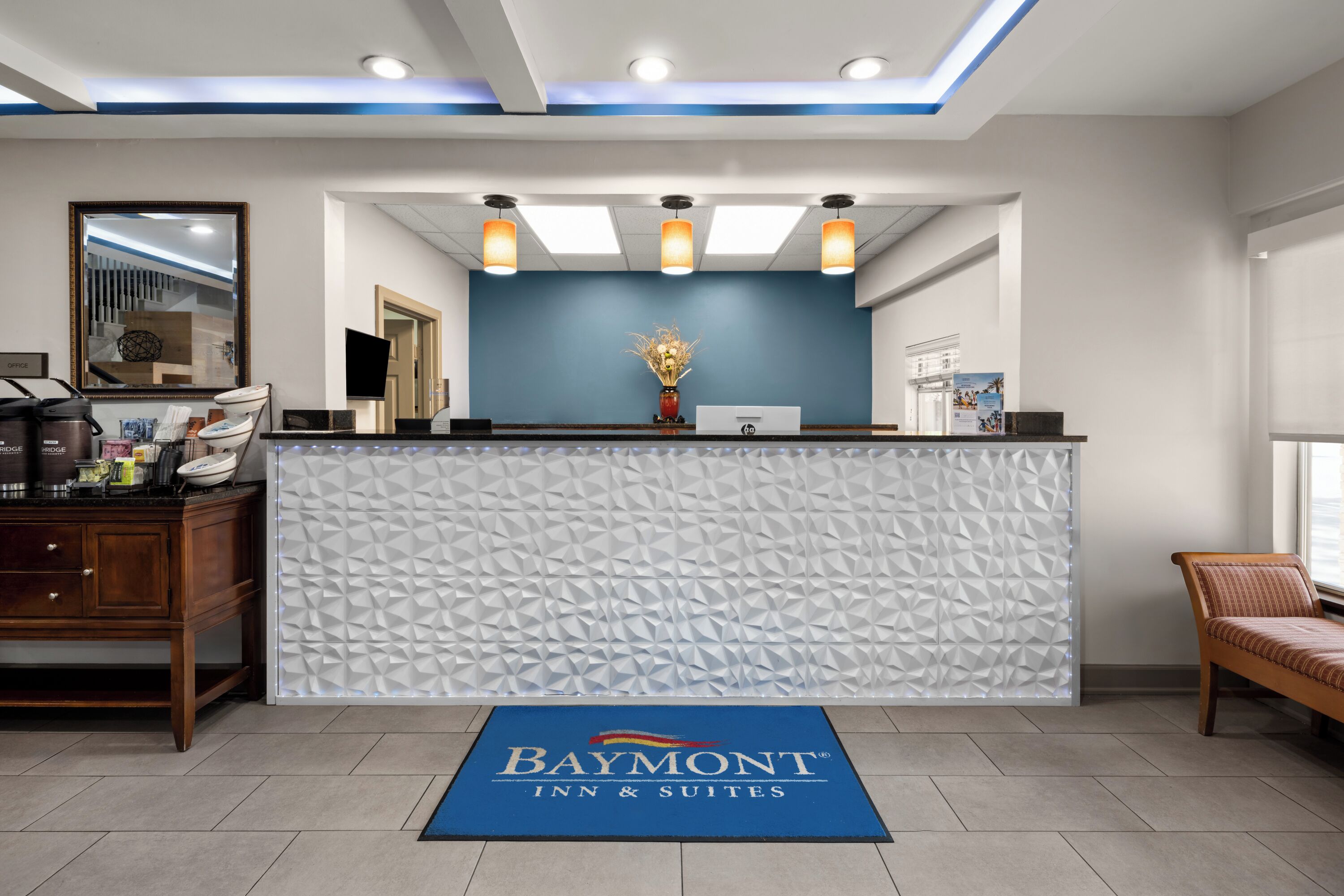 A reception area with a Baymont Inn & Suites sign, a reception desk, a wall-mounted TV, and a wooden cabinet with toiletries.