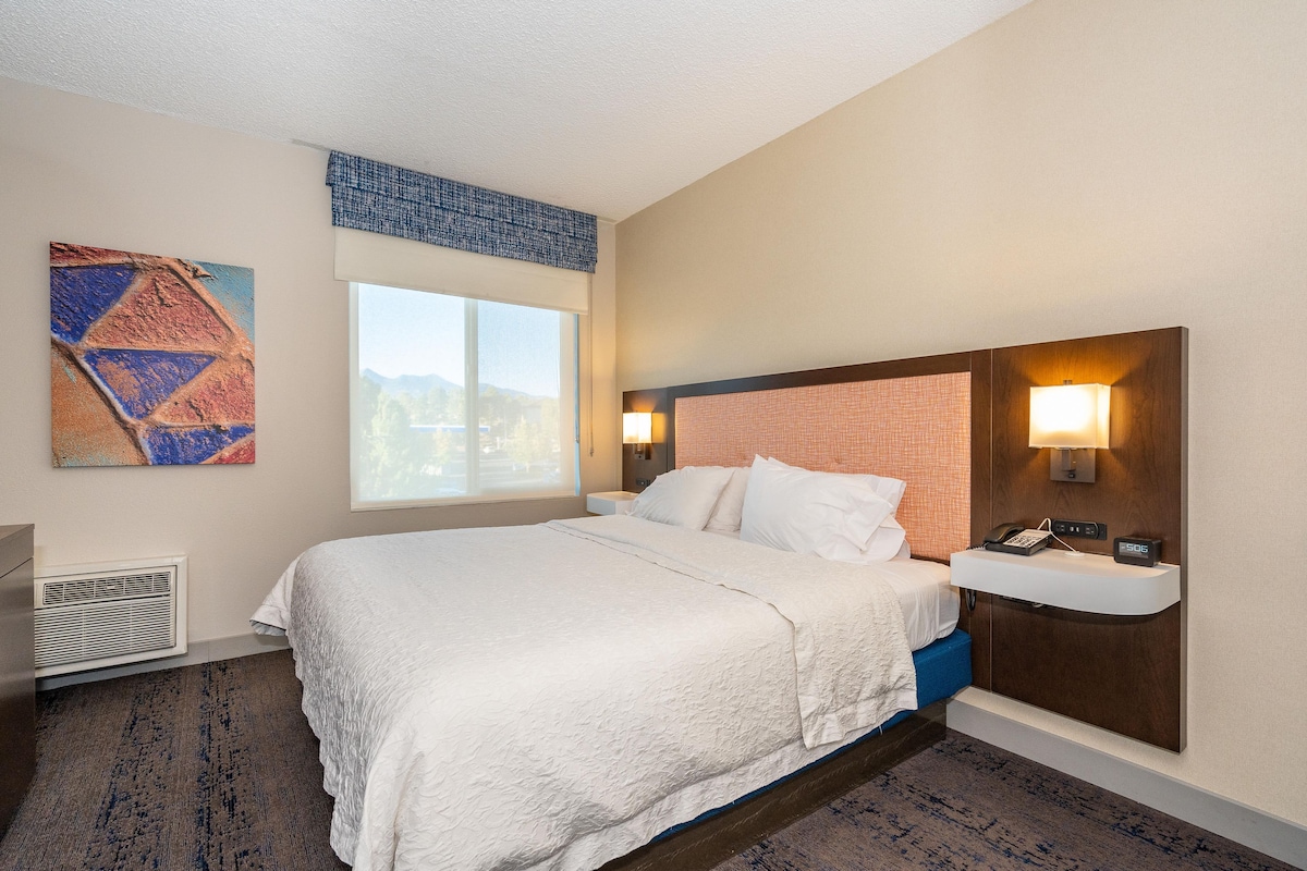 A hotel room with a bed, a nightstand, a lamp, a painting, and a window.