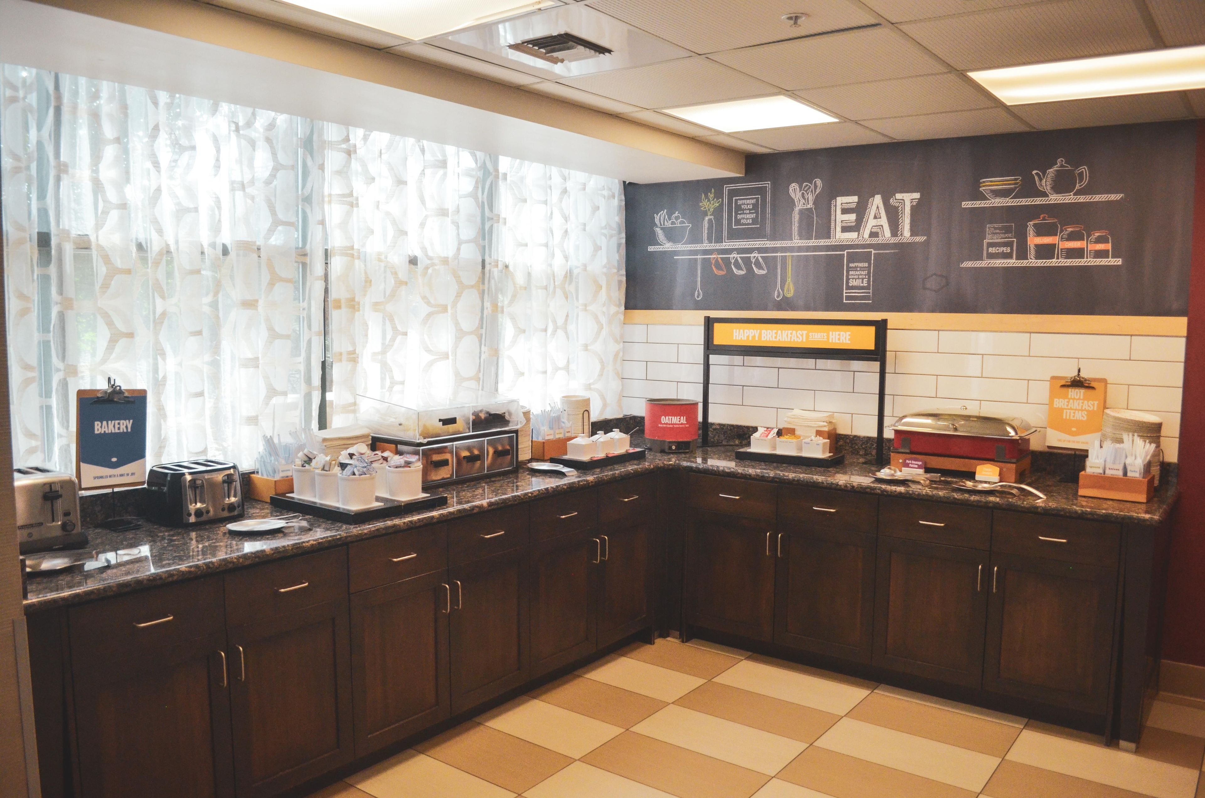 A breakfast area with a sign reading 'HAPPY BREAKFAST STARTS HERE,' a counter with various breakfast items, and a wall with a chalkboard menu.