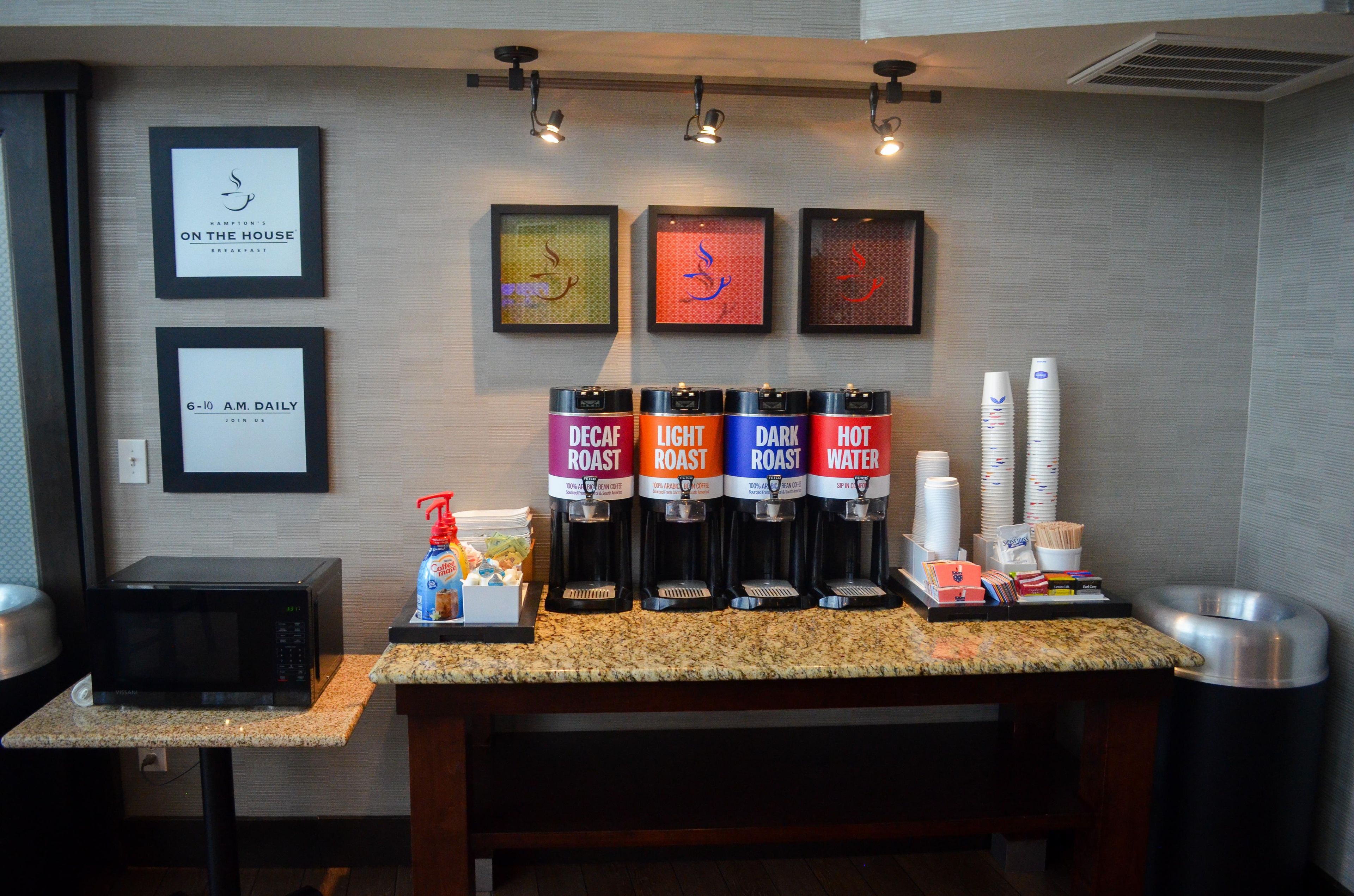 A coffee station with four coffee dispensers labeled Decaf Roast, Light Roast, Dark Roast, and Hot Water. There are framed signs on the wall and a microwave on the counter.