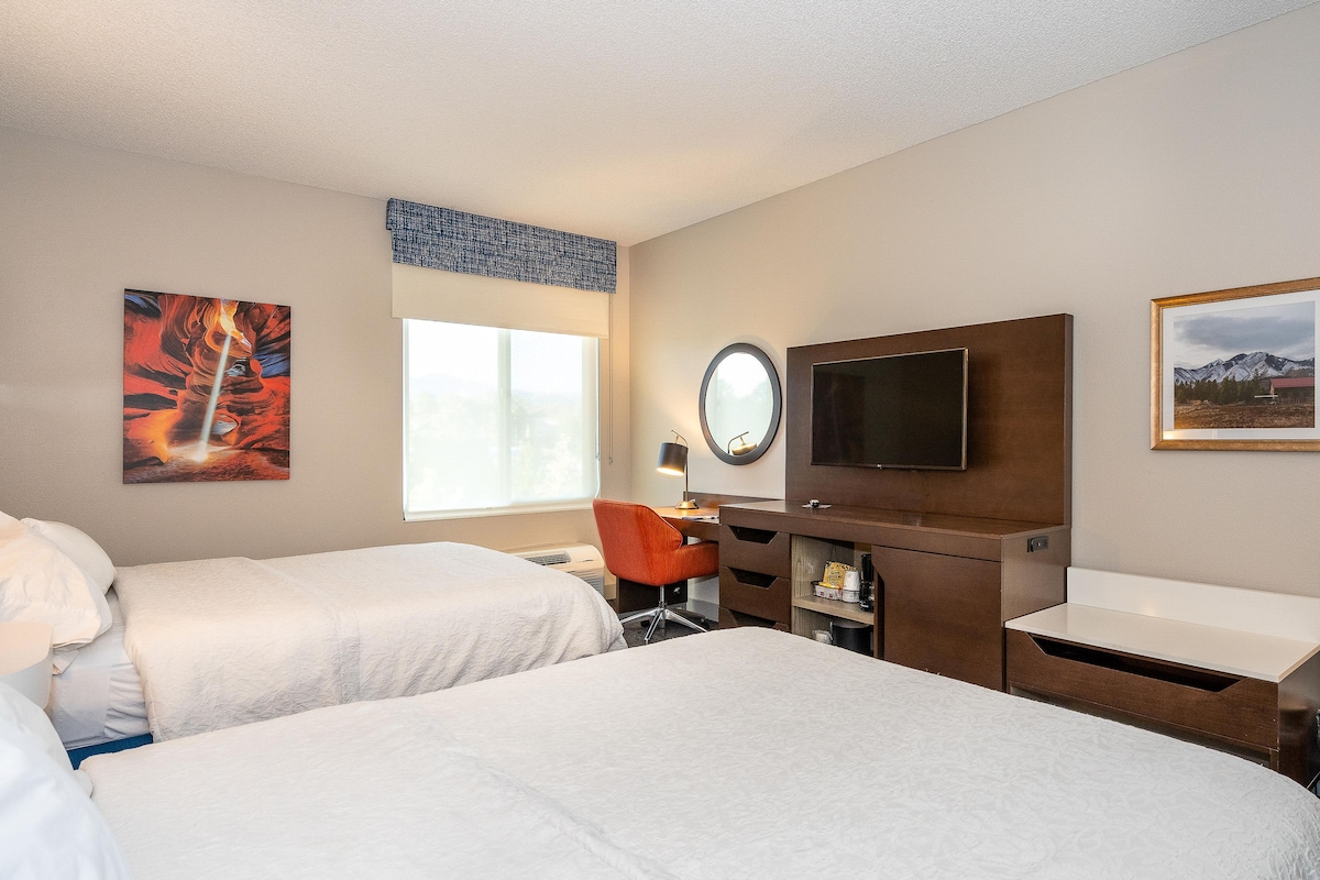 A hotel room with two beds, a desk, a chair, a TV, and artwork on the walls.