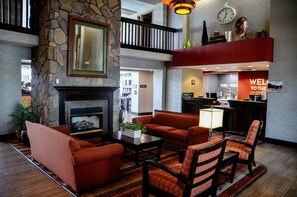 A hotel lobby with a stone fireplace, seating area, and a kitchen area with a 'Welcome' sign.