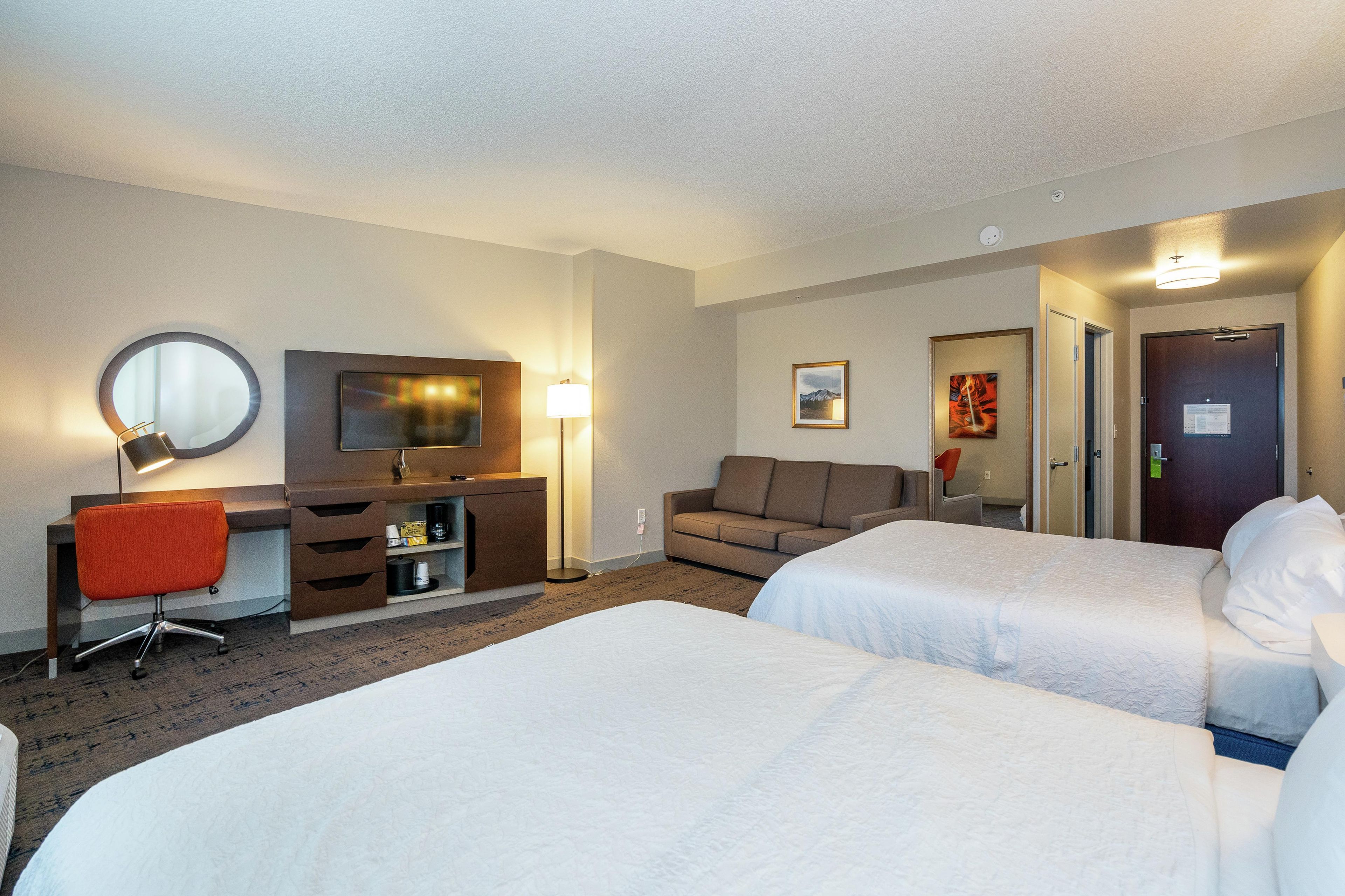 A hotel room with two beds, a desk with a chair, a sofa, a TV, and a mirror.