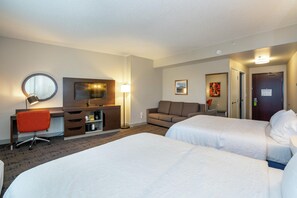 A hotel room with two beds, a desk with a chair, a sofa, a TV, and a mirror.