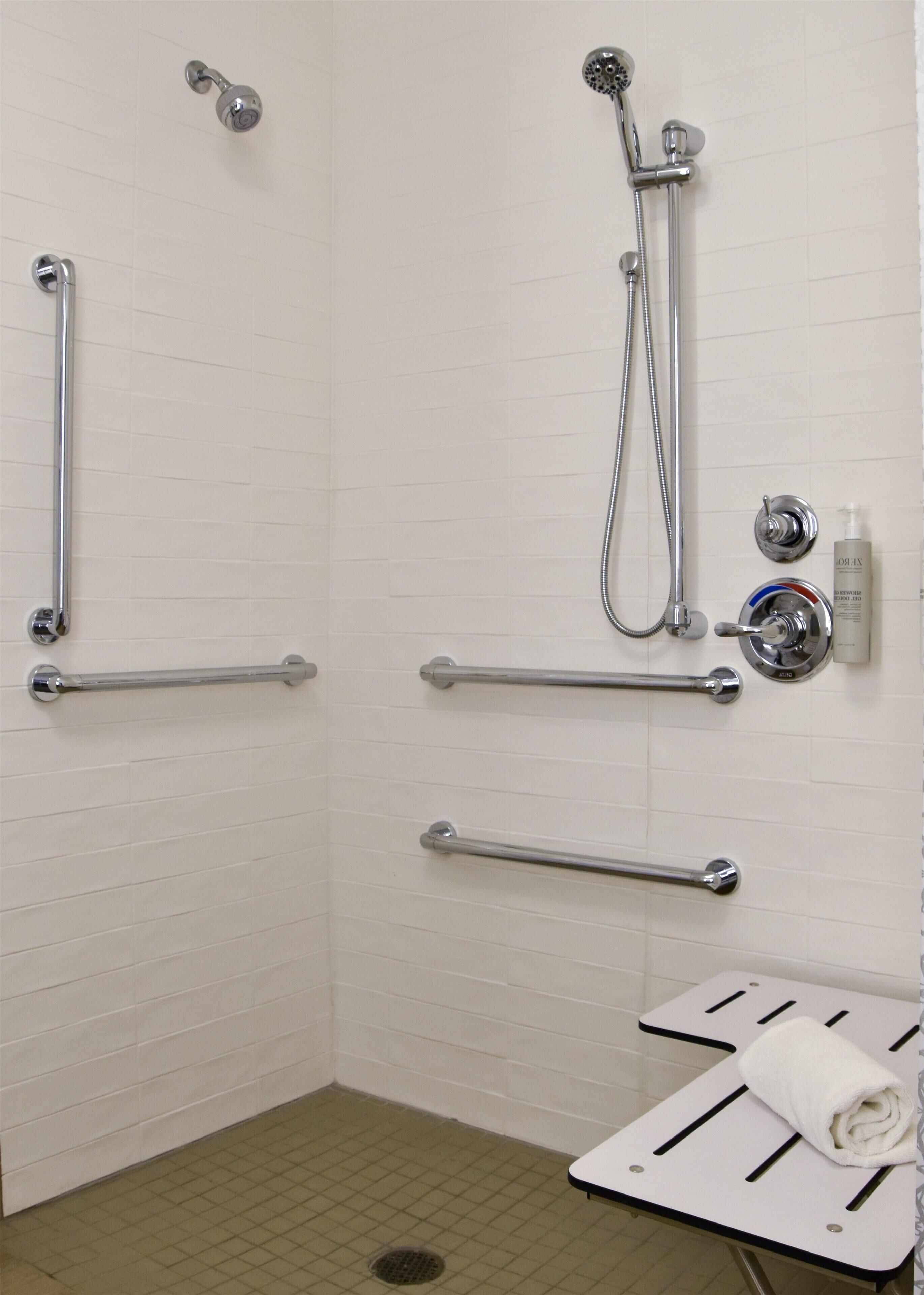 A bathroom with white tiled walls, a shower with a handheld showerhead, grab bars, a towel rack, and a bottle of hand soap.