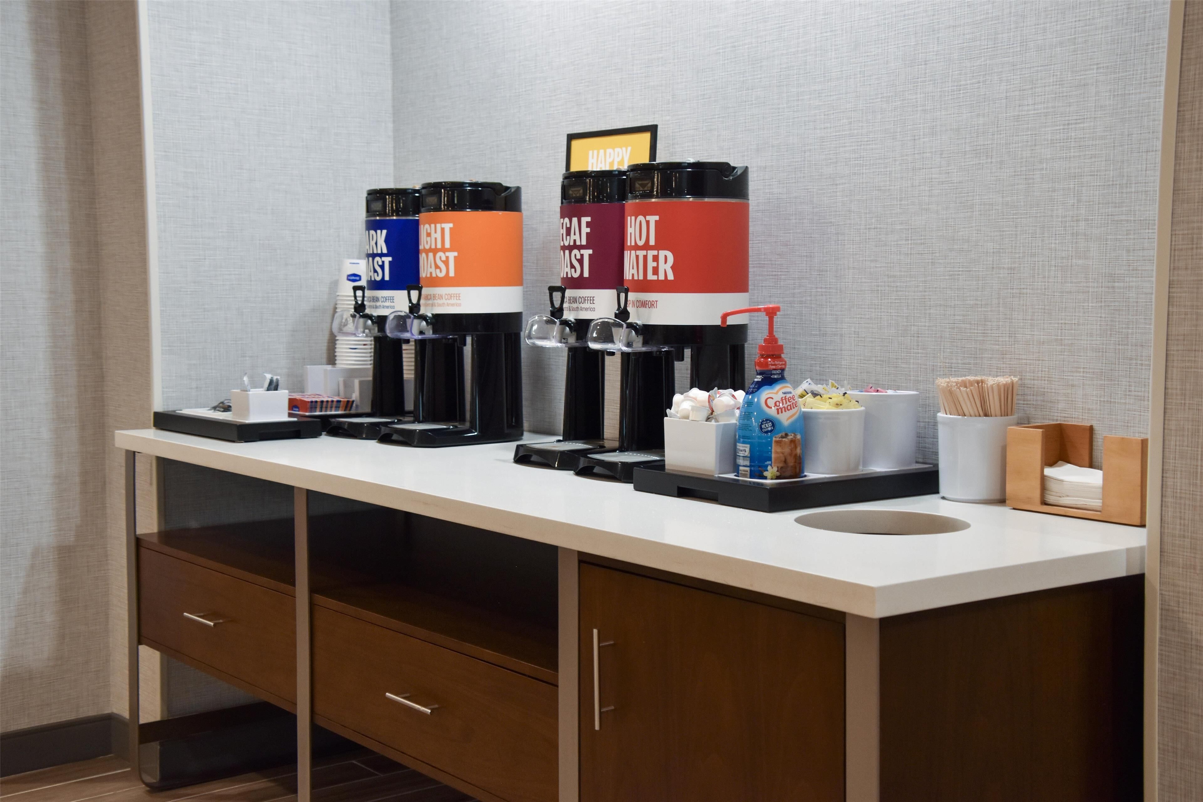A hotel room with a kitchenette featuring coffee machines labeled "Dark Roast" and "Hot Water," a coffee maker, a bottle of Coca-Cola, and a container of marshmallows.