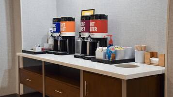 Habitación de hotel con kitchenette que incluye cafeteras etiquetadas como “Dark Roast” y “Agua Caliente”, una cafetera, una botella de Coca-Cola y un recipiente con malvaviscos.