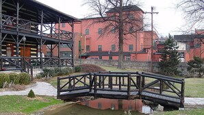 A wooden bridge over a small pond with a stone pathway leading to a building with a red brick facade and a metal structure in the foreground. - Hampton Inn Frankfort (Frankfort)