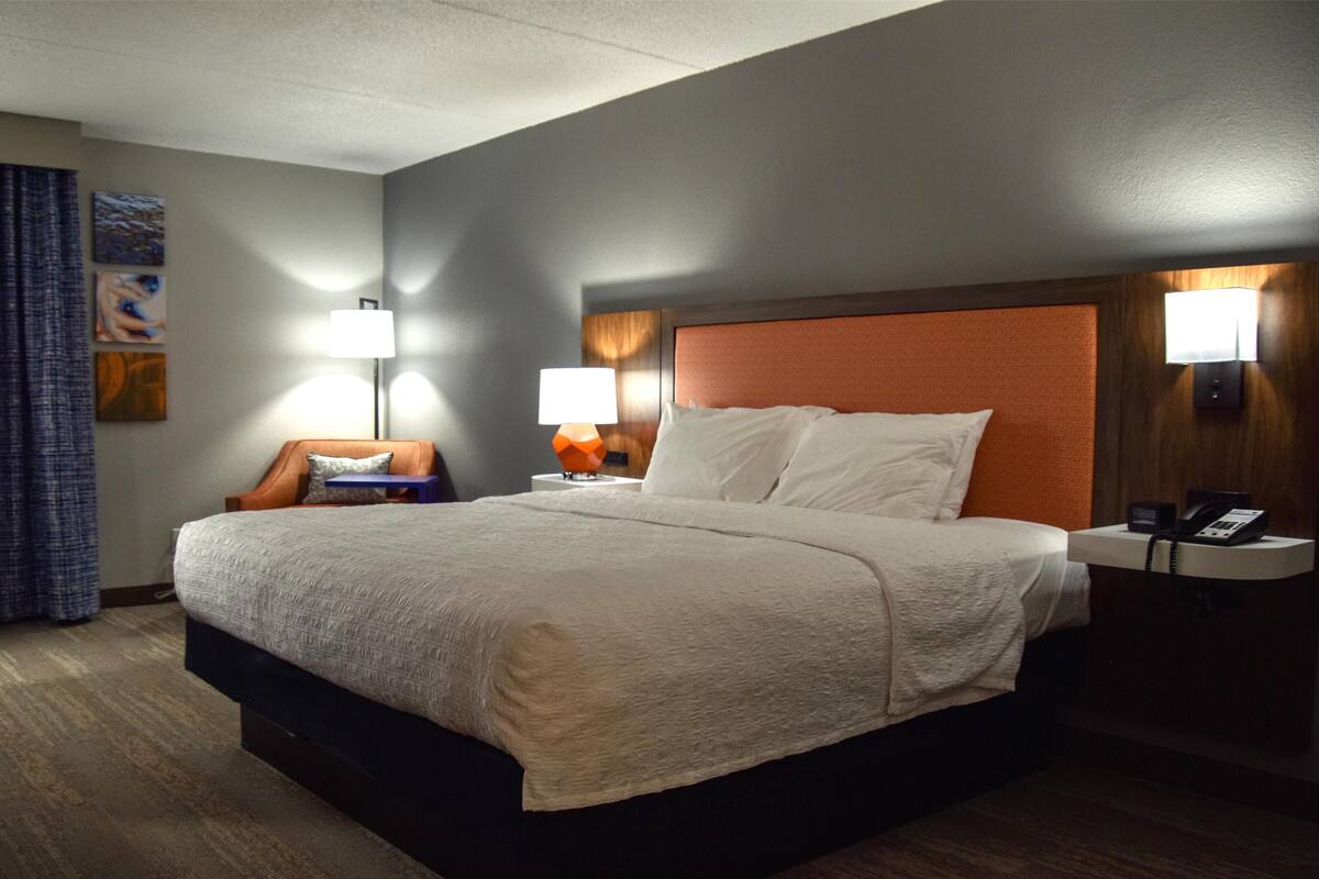Una camera d'albergo con un letto grande, due comodini con lampade, una sedia e un'opera d'arte appesa al muro.