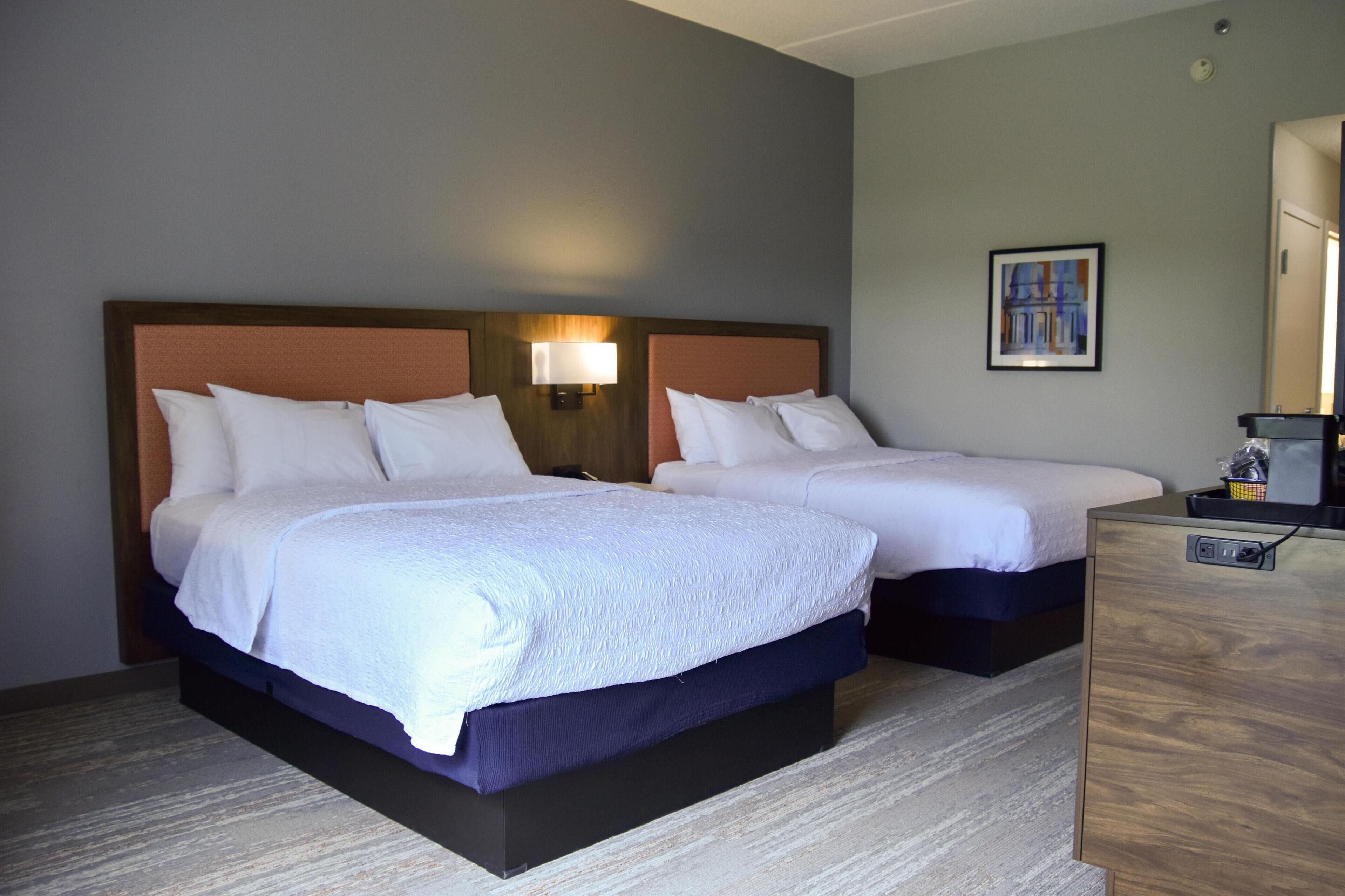 A hotel room with two beds, a nightstand, a lamp, and a painting on the wall.