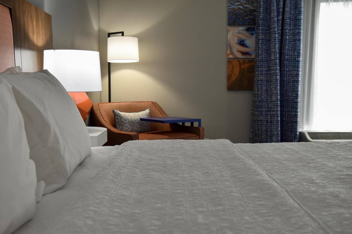 Una camera d'hotel con un grande letto, un comodino, una lampada, una sedia e un quadro appeso al muro.