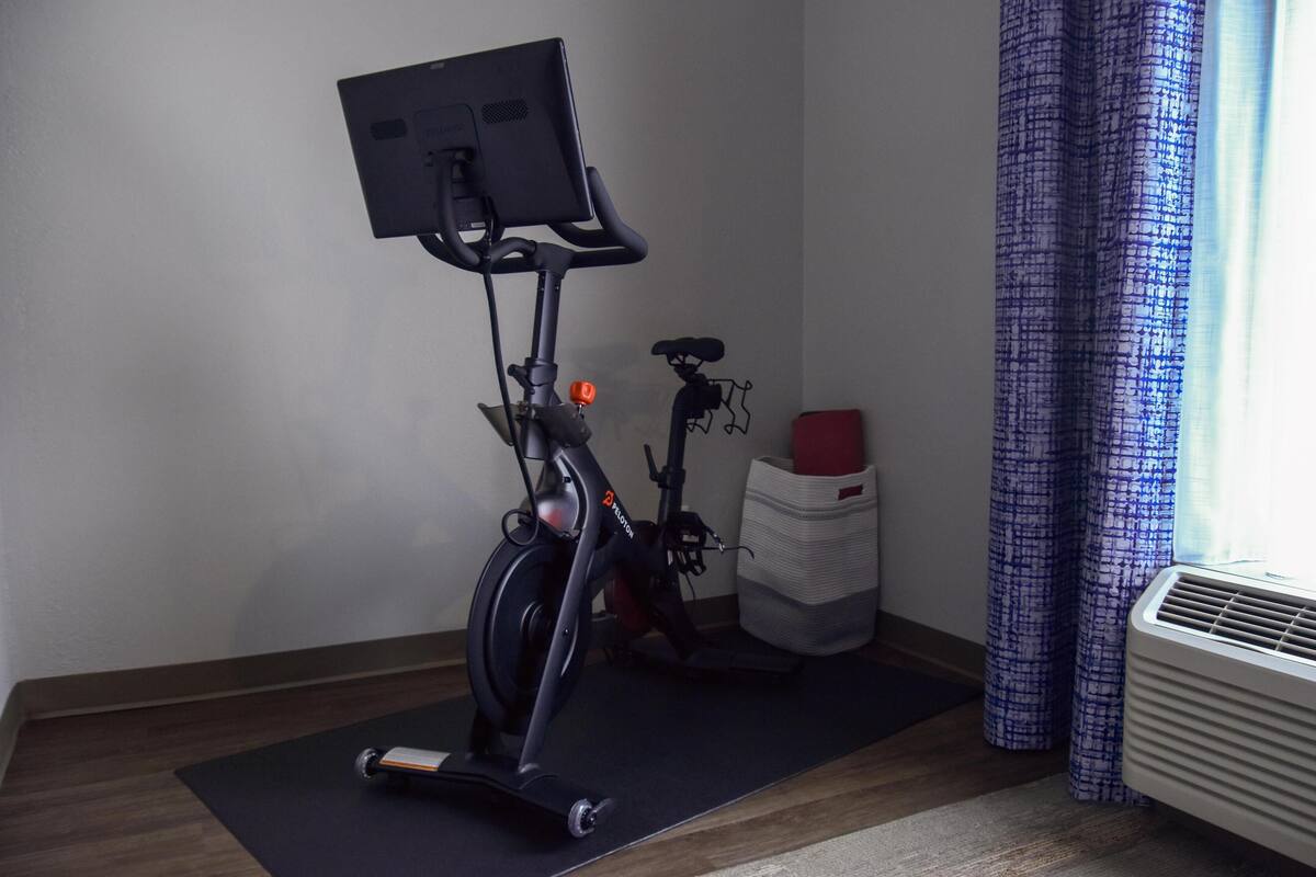 Una cyclette con schermo e sella rossa, un tavolino e una finestra con tende.