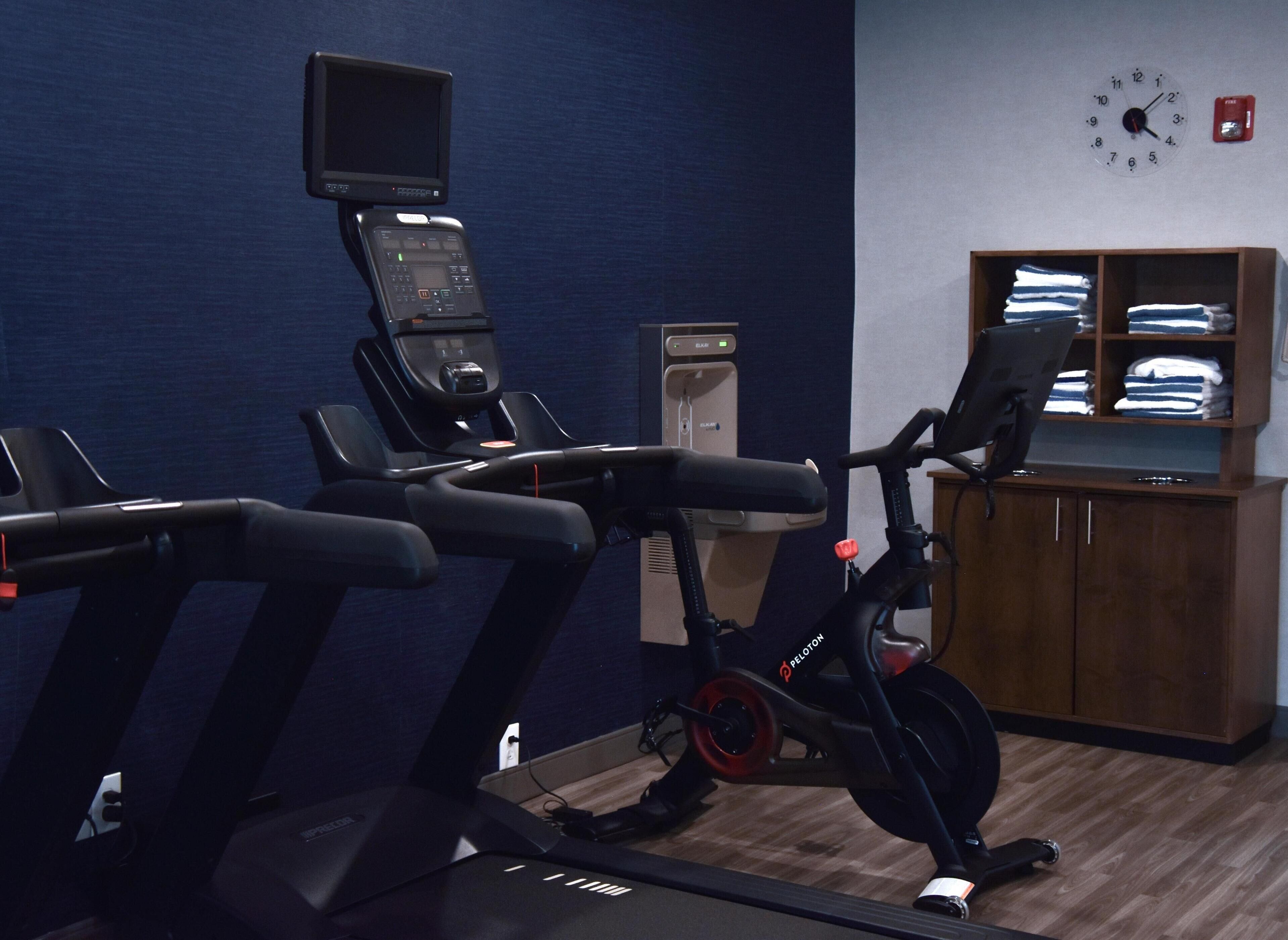 A gym with exercise equipment, a water dispenser, a clock, and neatly folded towels on shelves.