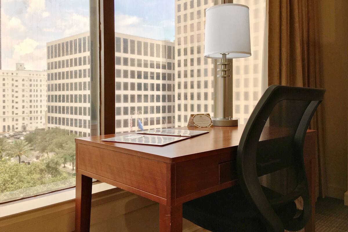 A hotel room with a wooden desk, a black office chair, a lamp, and a view of tall buildings through a window.